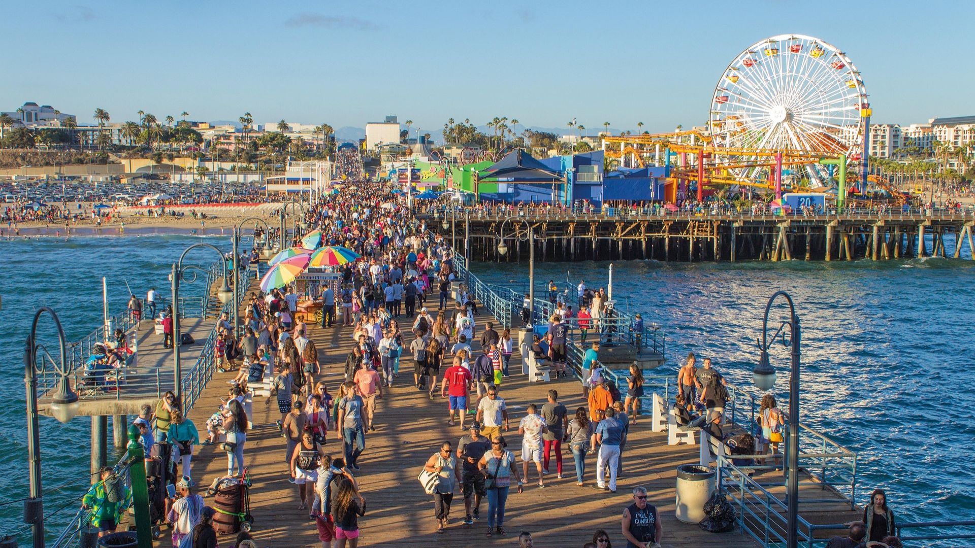 Santa Monica Pier with Ferris wheel and roller coaster rides at Pacific Park, bustling beach with people enjoying the waves and sand, and mountains in the background under a clear blue sky.