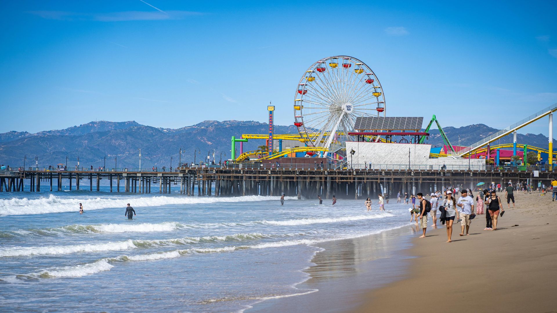 Santa Monica Pier with Ferris wheel and amusement park, viewed from the sandy beach with ocean waves. People can be seen walking along the shore and enjoying the sunny day.