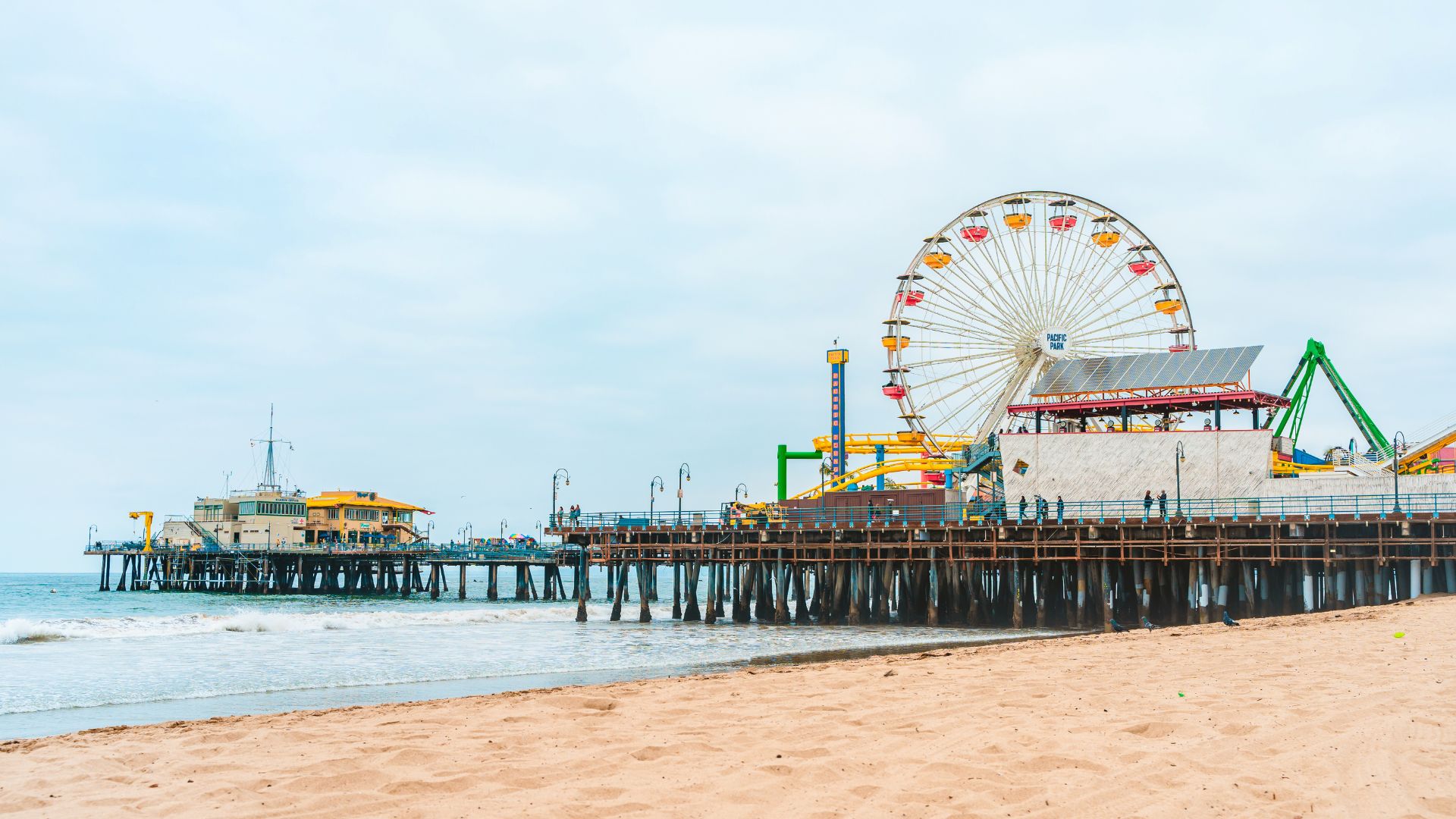 Santa Monica Pier with Ferris wheel and roller coaster rides at Pacific Park, bustling beach with people under a clear blue sky.