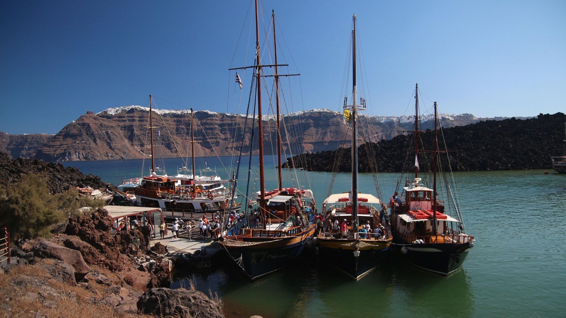 A harbor scene with boats docked against a volcanic cliff backdrop, in Santorini.