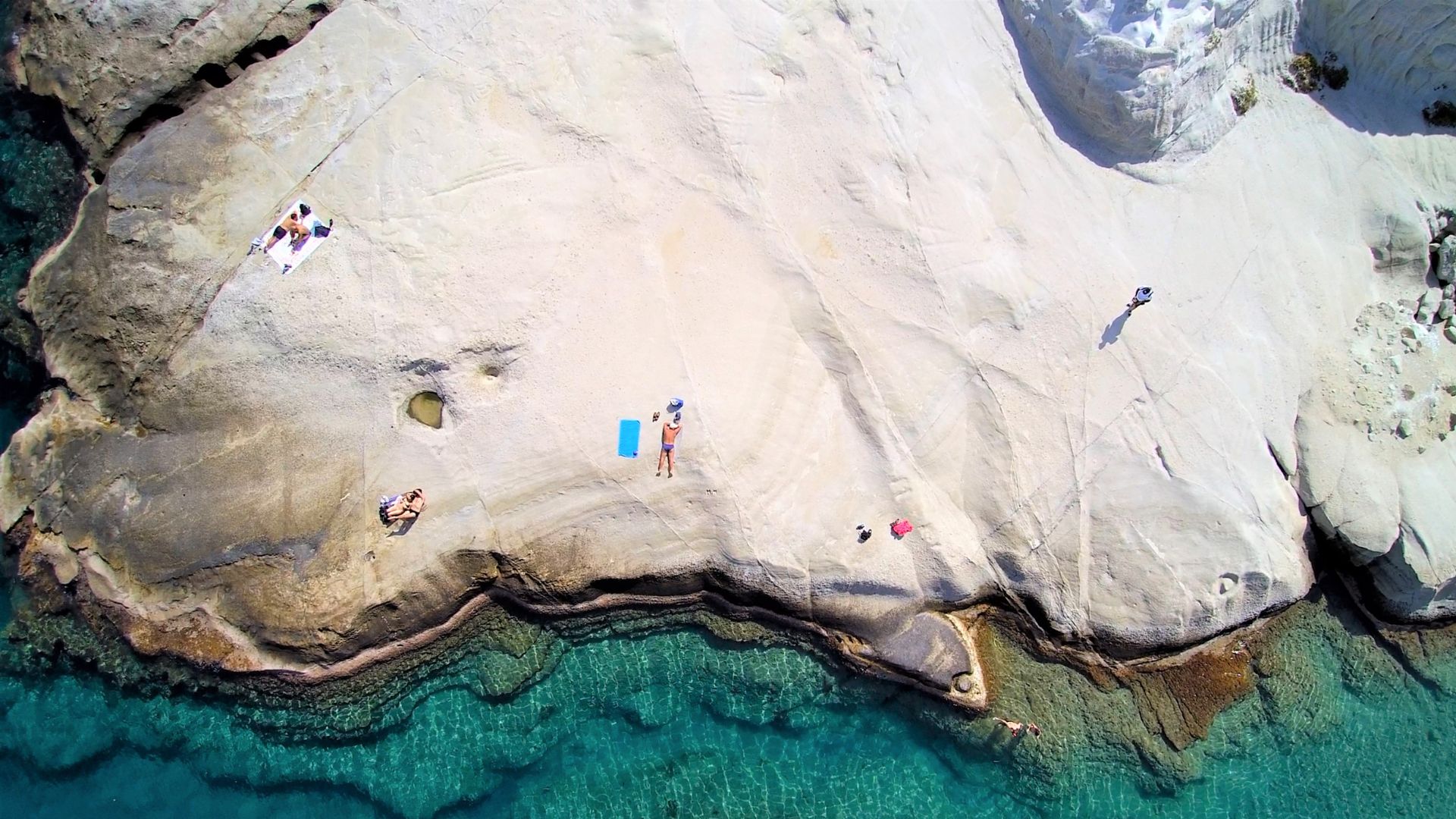 Sarakiniko Beach in Milos, Greece, features a unique lunar landscape of white volcanic rock formations against a backdrop of turquoise sea with beaachgoers at the rock.