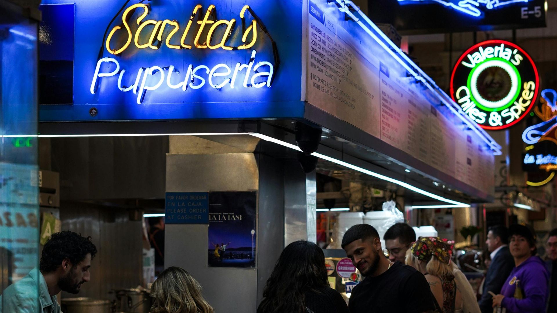 A vibrant indoor food court scene at night, featuring Sarita's Pupuseria with bright blue neon signs. People are seated at a counter and waiting in line, with other food stalls visible in the background under warm lighting.