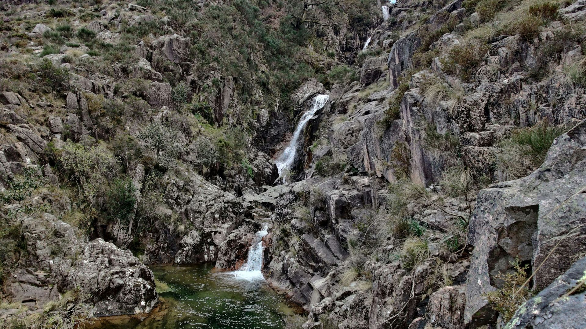A multi-tiered waterfall cascades into emerald-green natural pools surrounded by rocky, forested mountains under a cloudy sky in Peneda-Gerês National Park, Portugal.