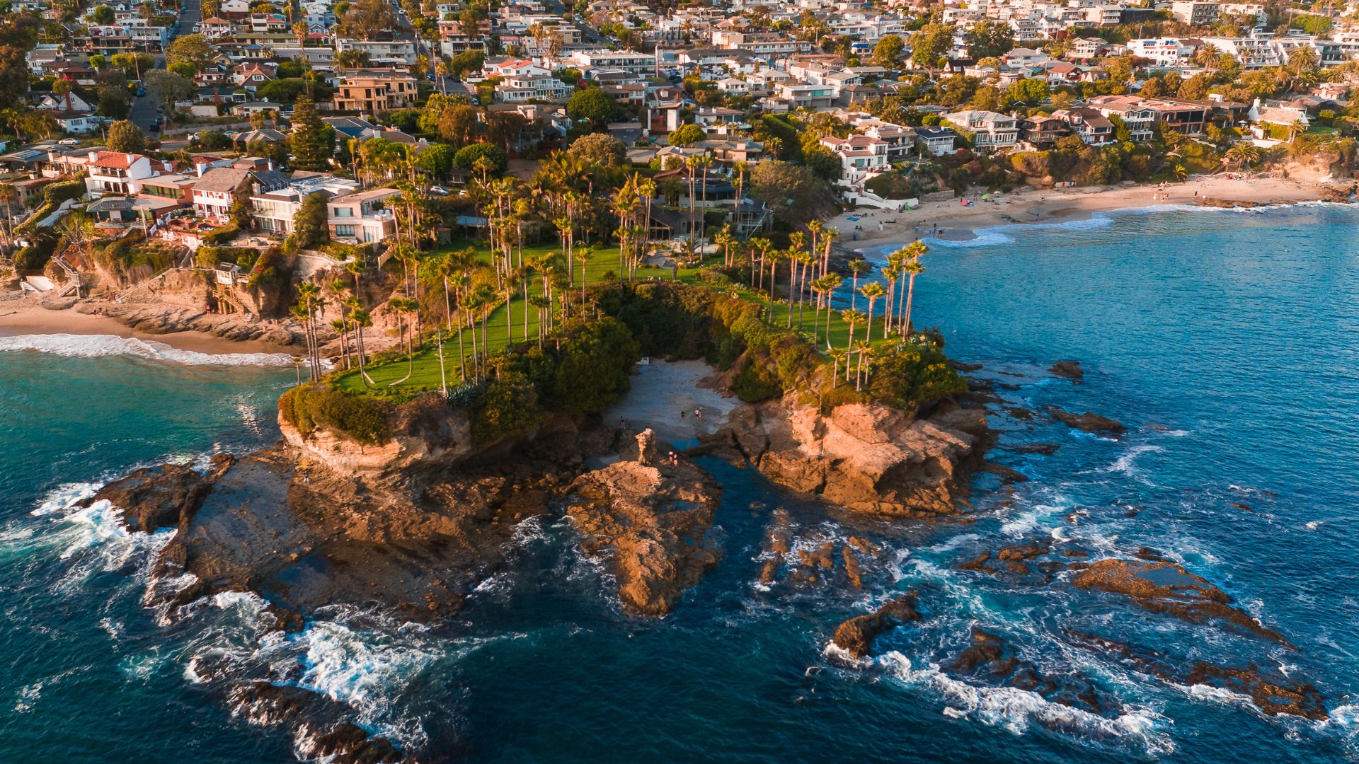 An aerial view of Shaw's Cove in Laguna Beach, California, featuring a lush green headland with palm trees extending into the clear blue ocean, surrounded by rocky shorelines and residential buildings nestled on the hills above.