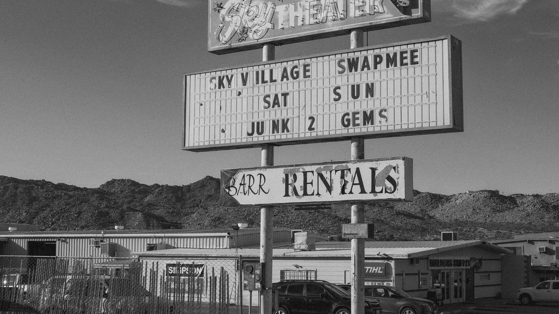 A black and white photograph of the large, multi-panel sign for the Sky Village Swap Meet and Sky Drive-In Theatre in Yucca Valley, California, with "BARR RENTALS" also visible. The sign stands tall against a cloudy sky and mountains in the background, with cars parked below and buildings visible behind a fence.