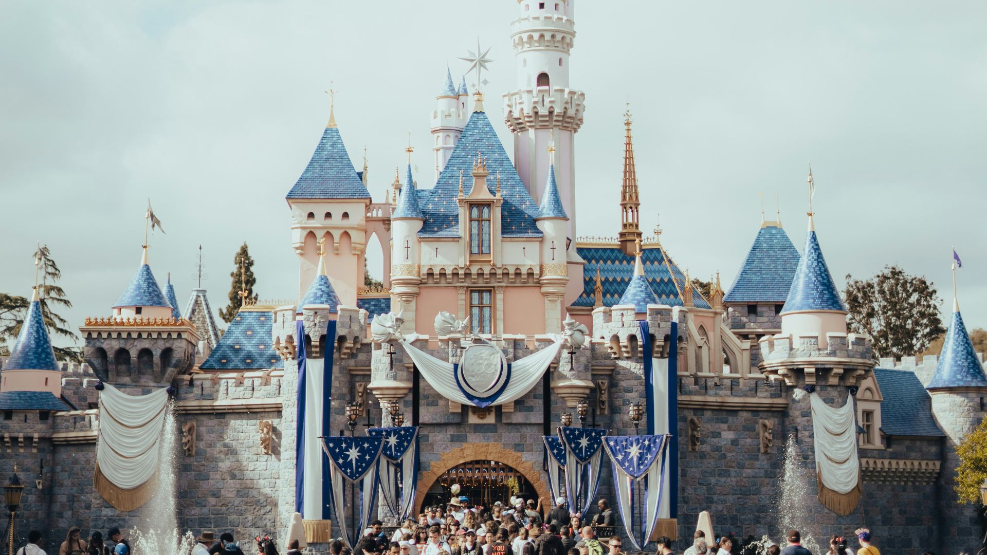 Sleeping Beauty Castle at Disneyland Park in Anaheim, California, with a crowd of visitors in front on a cloudy day.