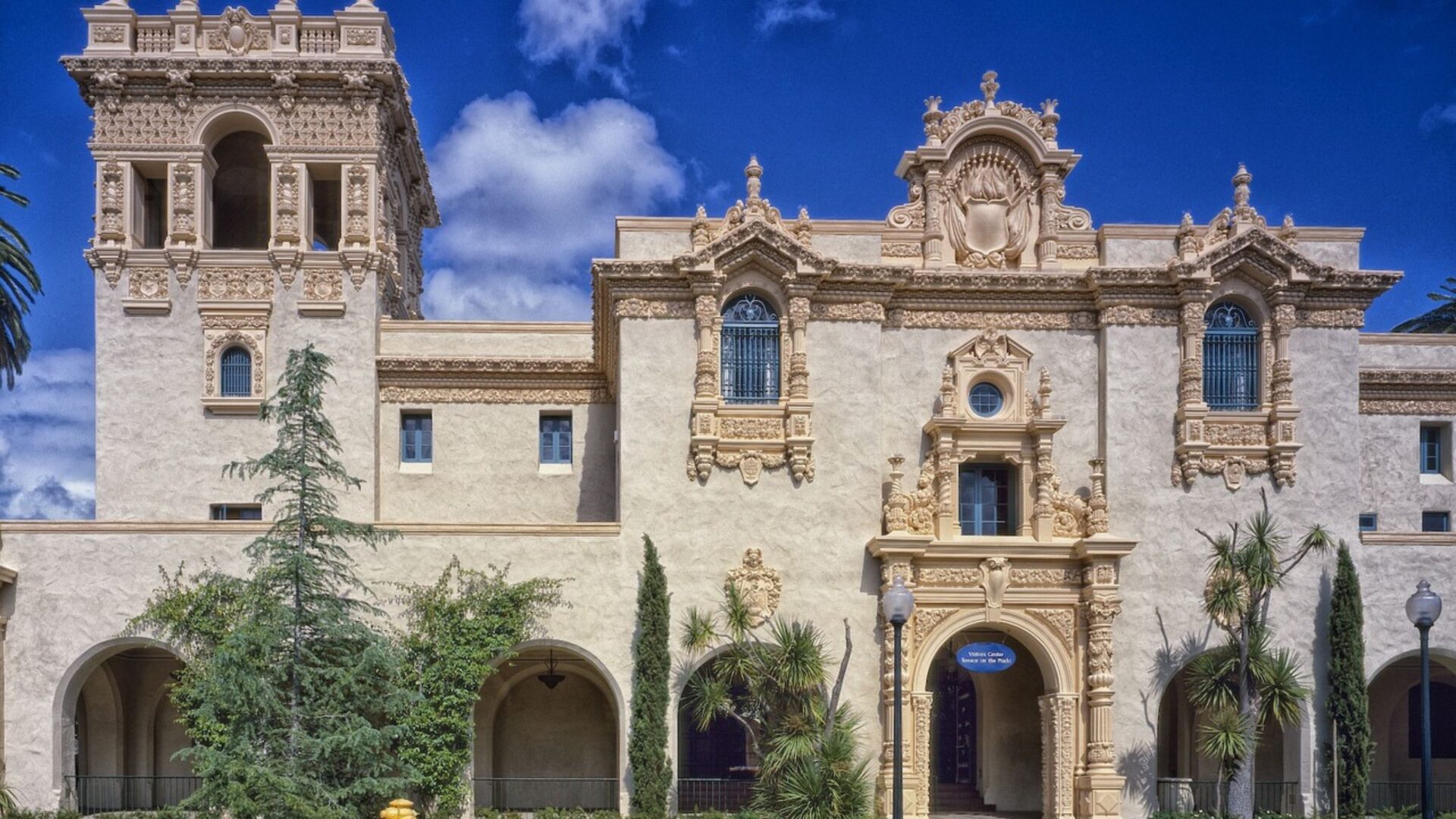 A grand, ornate building in the Spanish Colonial Revival style, featuring a tall tower on the left and intricate architectural details on the facade, set against a bright blue sky with scattered clouds. Green trees and shrubs are visible in front of the building.