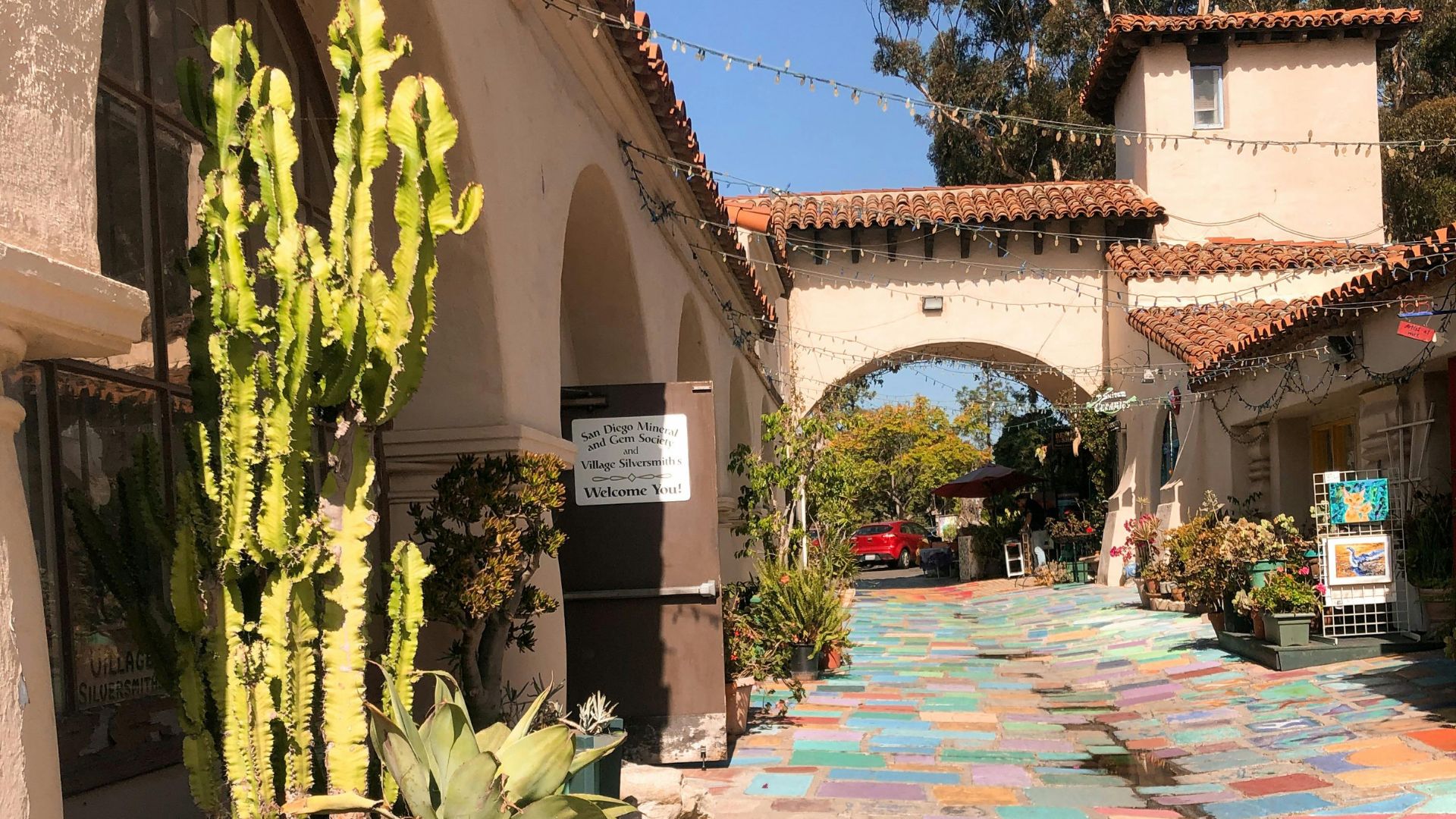 A vibrant outdoor scene at the Spanish Village Art Center in San Diego, California, showing colorful tiled pathways, arched doorways, and Spanish-style architecture, with a large cactus in the foreground and a red car visible in the distance through an archway.