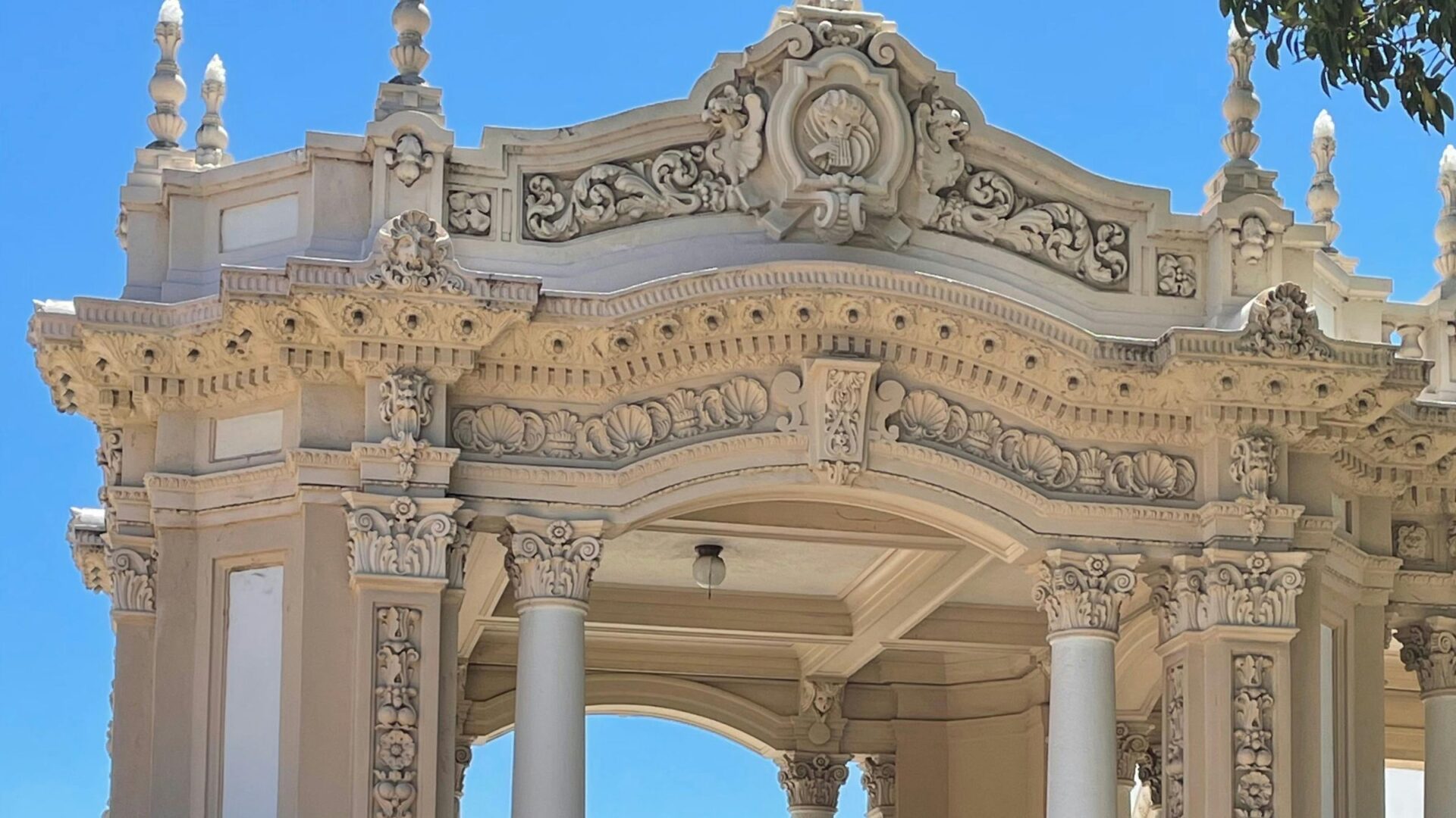 An ornate, light-colored stone pavilion with classical columns and intricate carvings, set against a clear blue sky, surrounded by green shrubs in Balboa Park, San Diego.