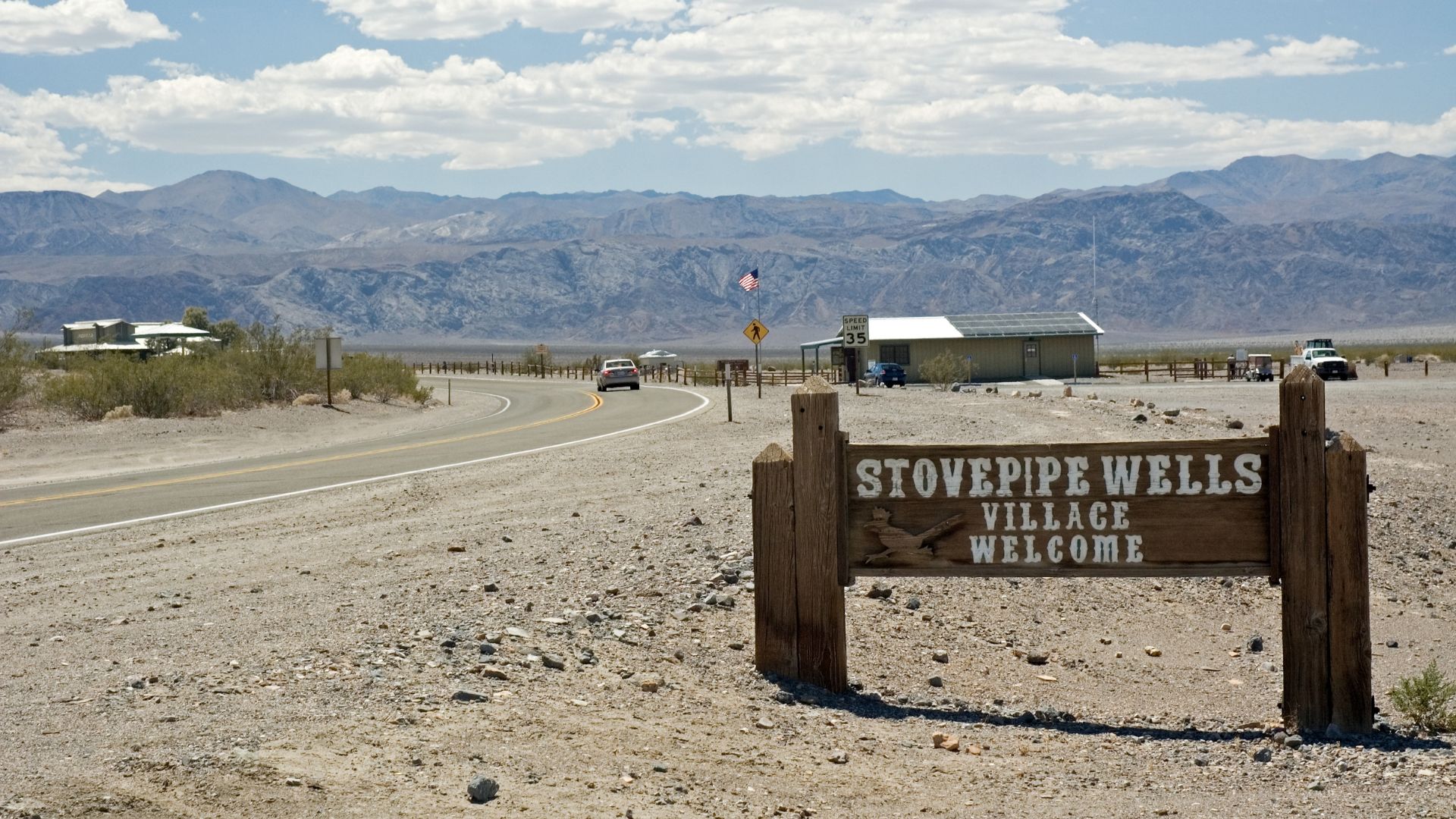 A wooden welcome sign for "STOVEPIPE WELLS VILLAGE" stands on the right side of a paved road leading into a desert landscape with buildings and mountains in the background under a partly cloudy sky.