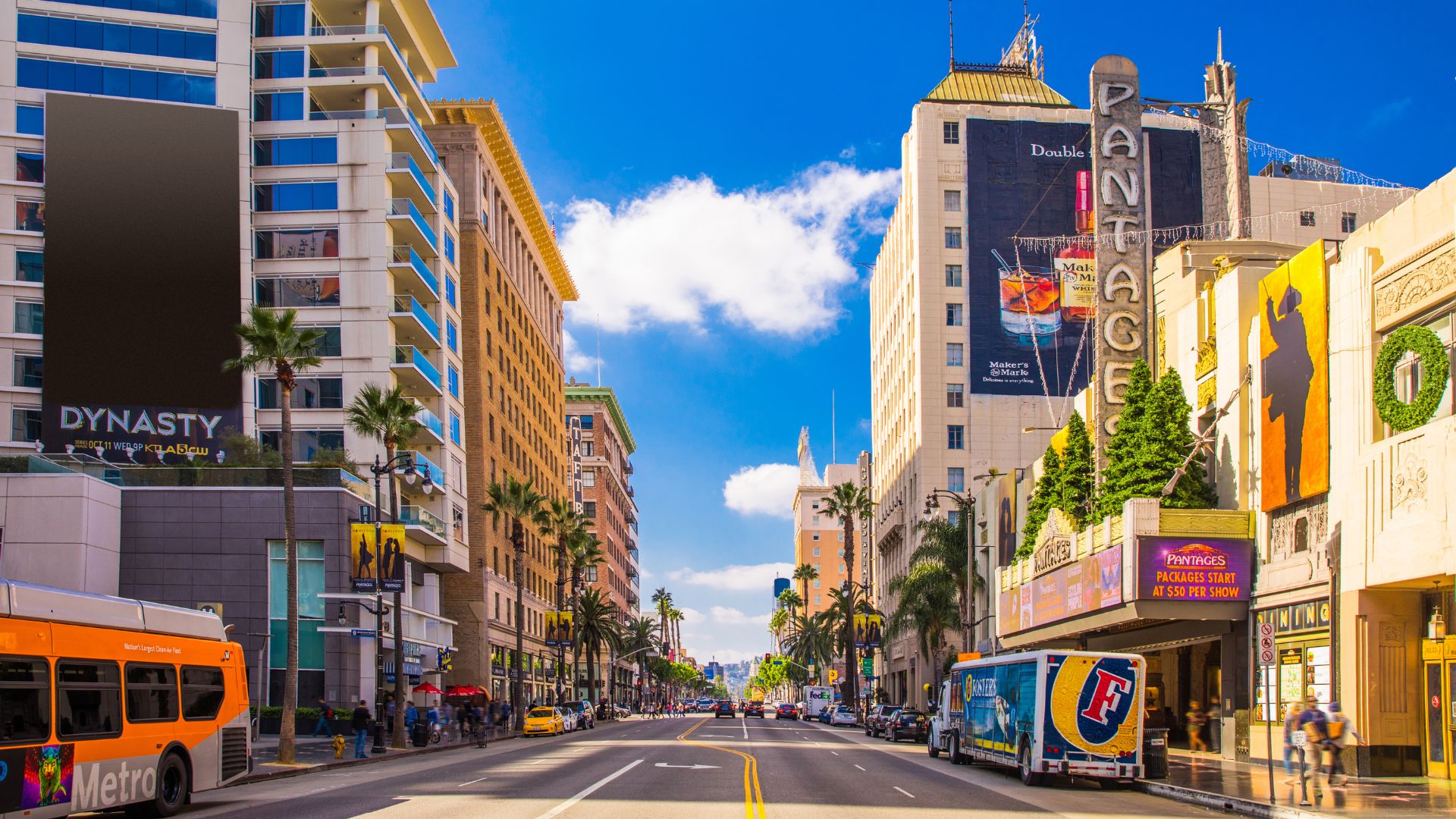 A daytime view of Sunset Boulevard in Los Angeles, California, showcasing a bustling street with tall buildings, palm trees, and a clear blue sky. The iconic Hollywood Pantages Theatre is visible on the right, with the 'DYNASTY' sign on a building to the left. A Metro bus and other vehicles can be seen on the road, highlighting the vibrant urban environment.