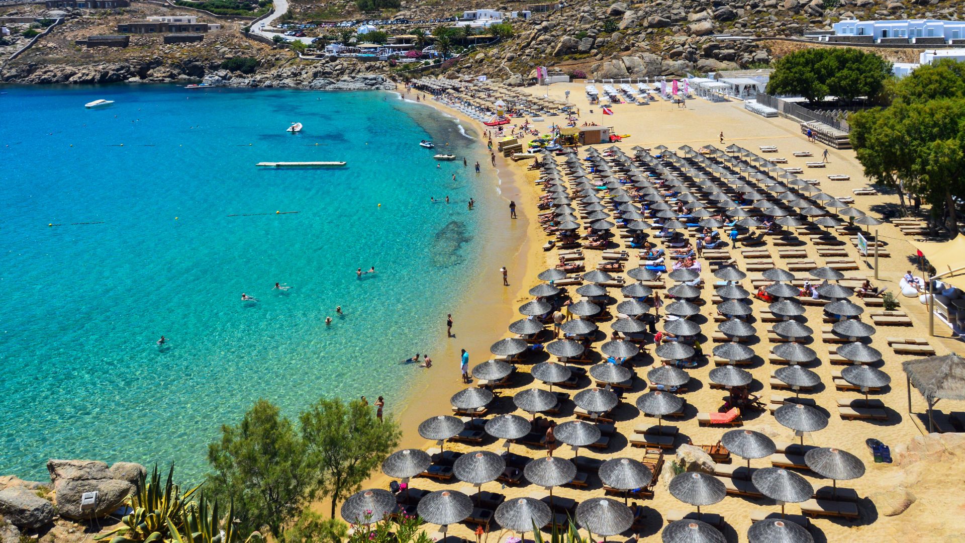 A high-angle view of Super Paradise Beach Club in Mykonos, Greece, with turquoise water, sandy beach, and beach umbrellas.