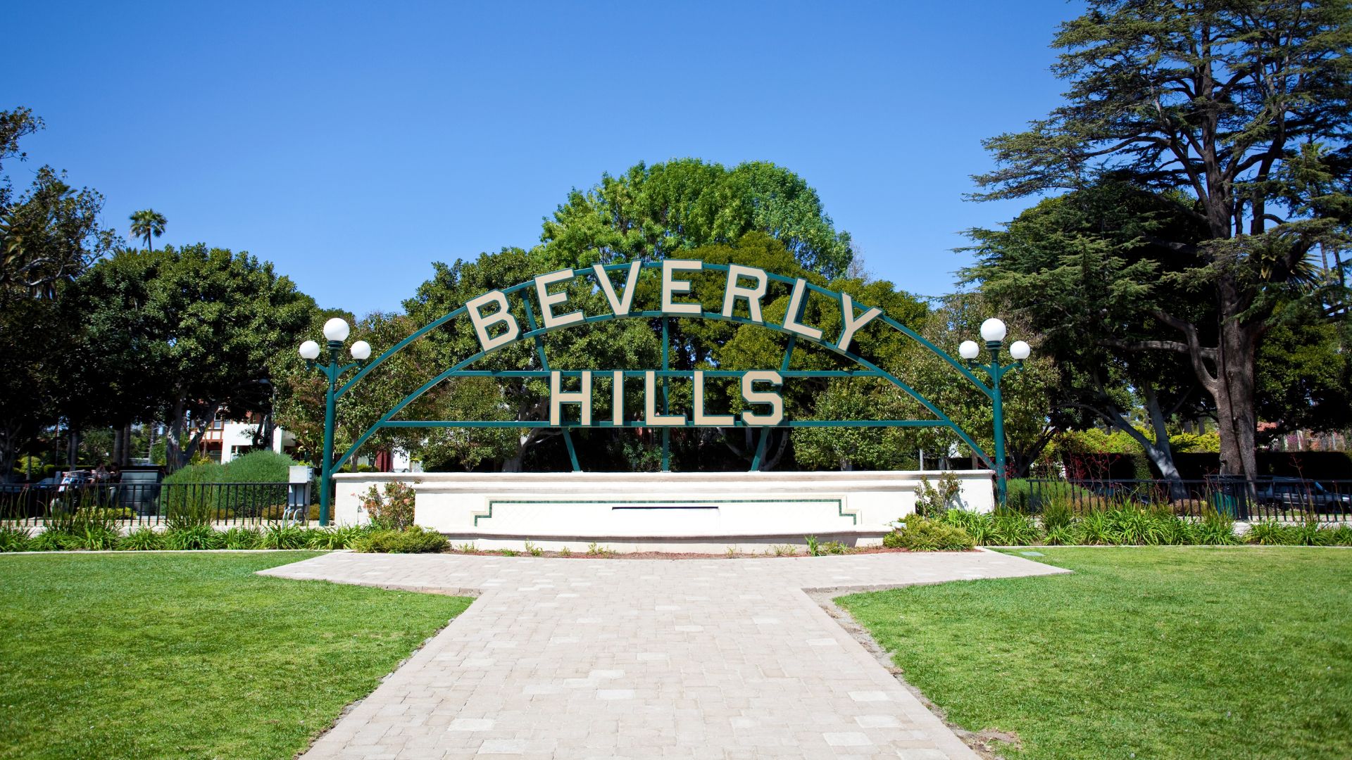 The iconic Beverly Hills sign, a green arch with white lettering, stands proudly in Beverly Gardens Park. A paved pathway leads to the sign, surrounded by lush green grass and trees under a clear blue sky.