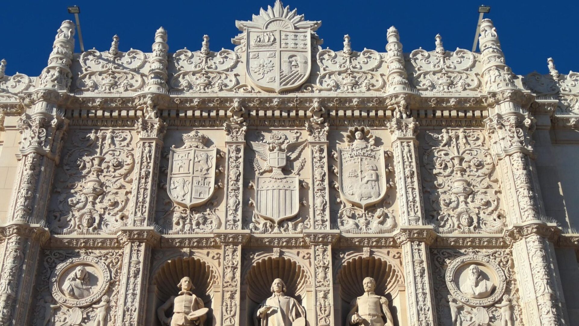 A close-up view of the highly ornate, light-colored stone facade of The San Diego Museum of Art, featuring intricate carvings, multiple coats of arms, and three statues of historical figures in recessed niches.
