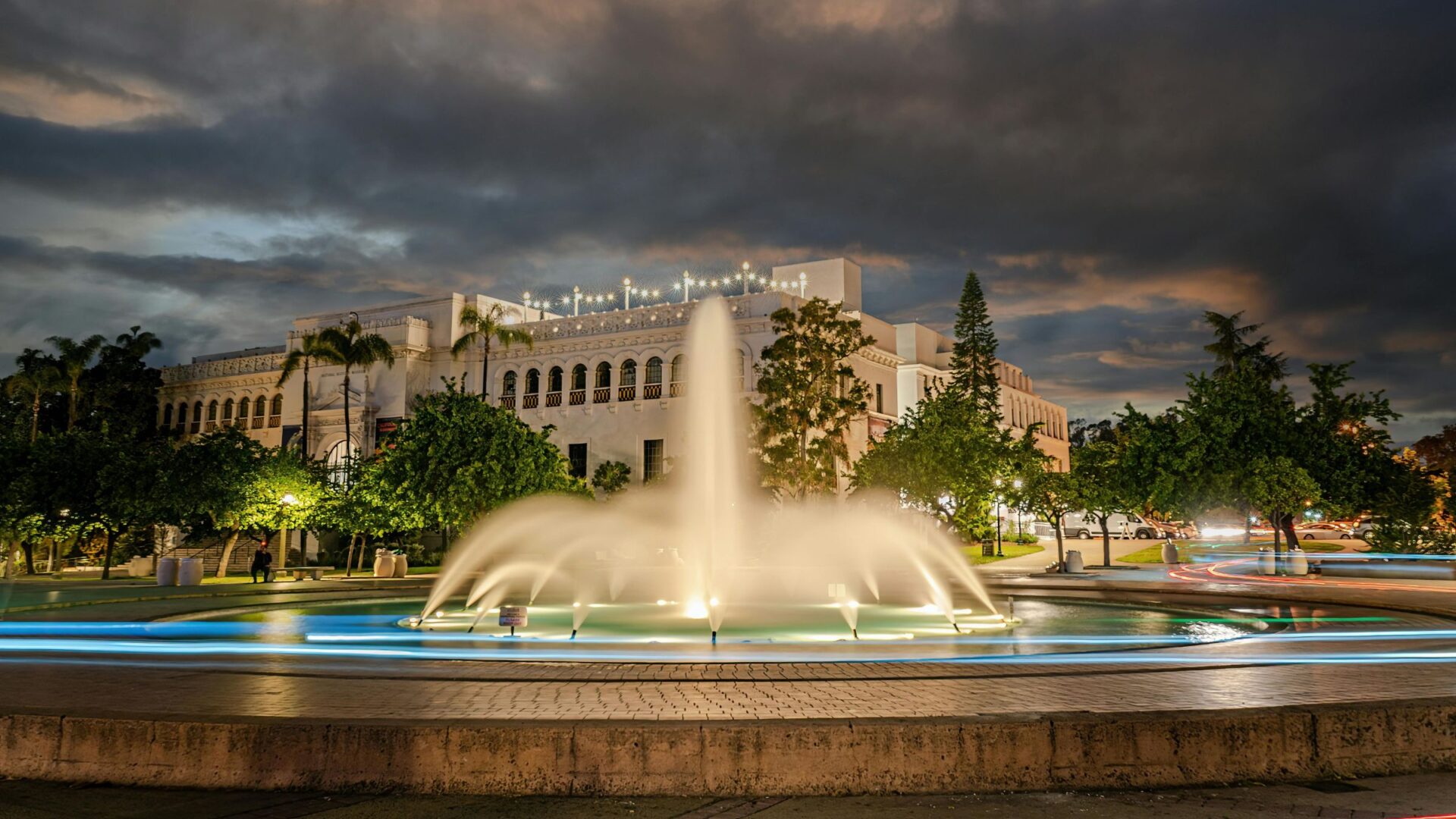 A long exposure photograph at dusk shows the Bea Evenson Fountain in the foreground with illuminated water streams, and the San Diego Natural History Museum building in the background under a cloudy sky in Balboa Park.
