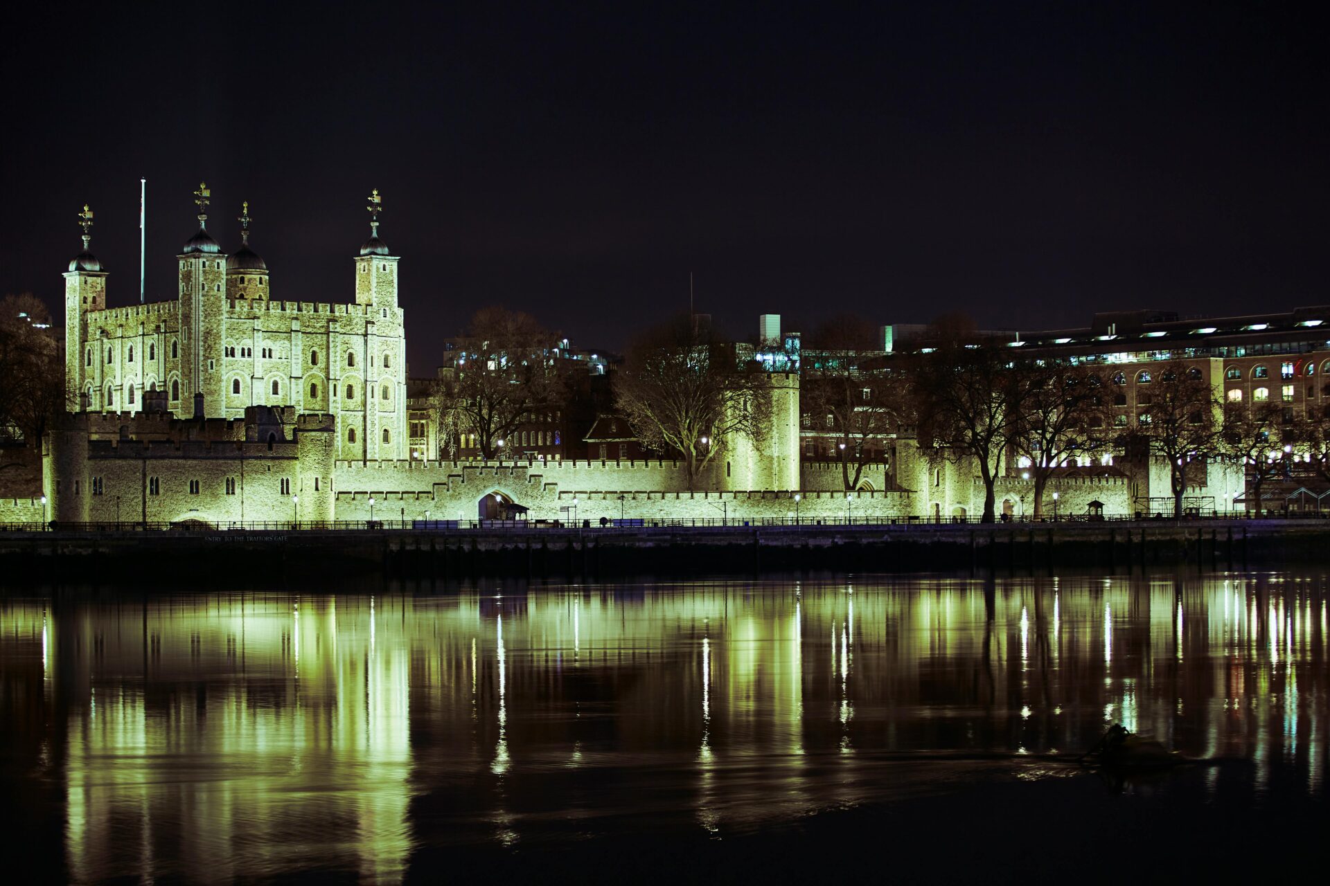 The Tower of London glowing under spotlights at night, its historic stone walls casting long shadows across the Thames River.