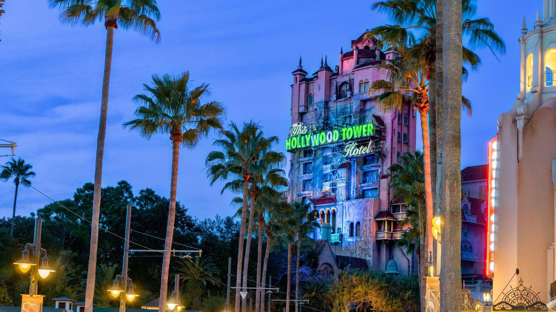 The Twilight Zone Tower of Terror at Disney's Hollywood Studios in Orlando, Florida, featuring its iconic hotel facade and neon sign at dusk. The palm trees add to the Florida setting. 