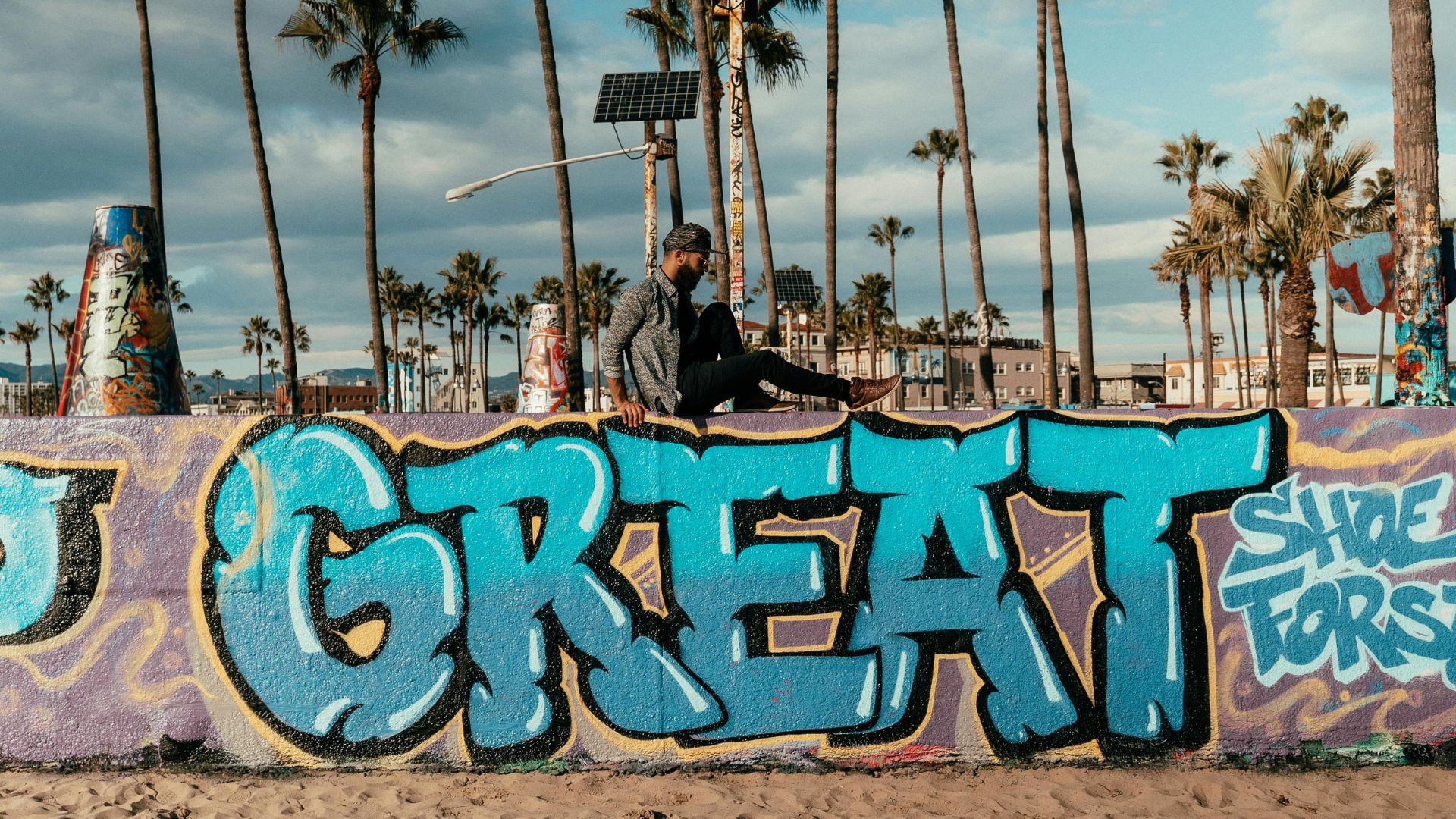 Man sits on graffiti-covered wall at Venice Art Walls, Venice Beach, California, with palm trees and blue sky background, featuring the word 'GREAT' in blue graffiti.