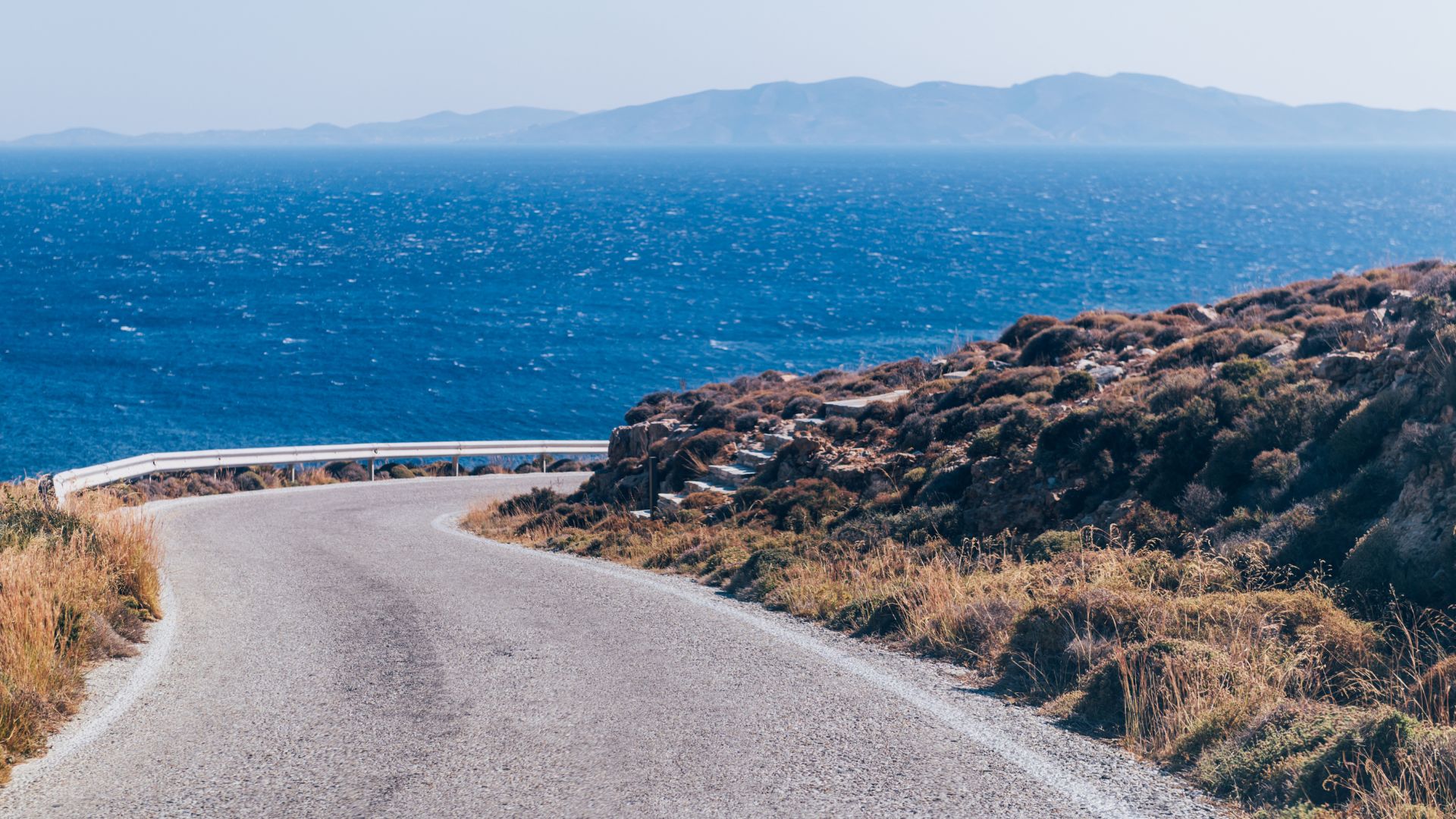 Winding coastal road in Tinos, Greece, with the Aegean Sea in the background.