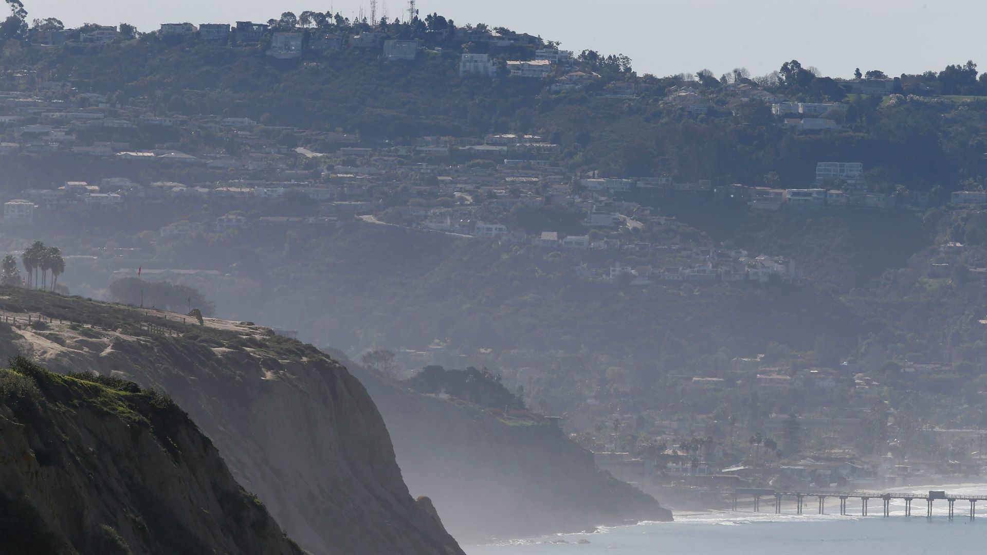 A wide shot of the coastline at Torrey Pines State Natural Reserve, featuring steep, sandy cliffs in the foreground, a hazy view of a distant pier and the Pacific Ocean, and a hillside covered with houses and trees in the background under a clear sky.