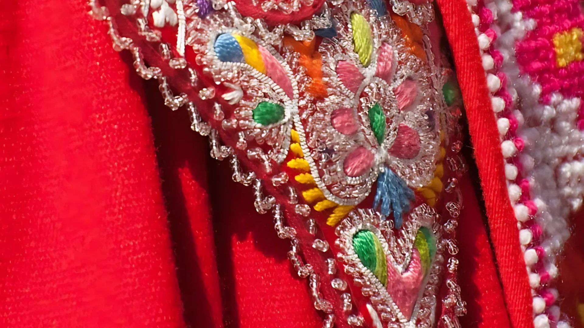 Close-up of richly embroidered red fabric, part of a traditional costume, featuring colorful floral patterns and silver accents.