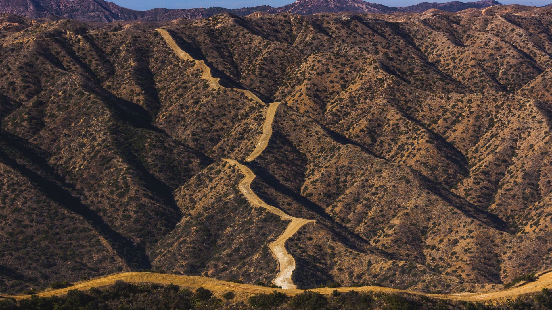 A winding dirt trail traverses a steep, arid mountainside on Catalina Island, California, with undulating ridges and sparse vegetation under a clear sky.