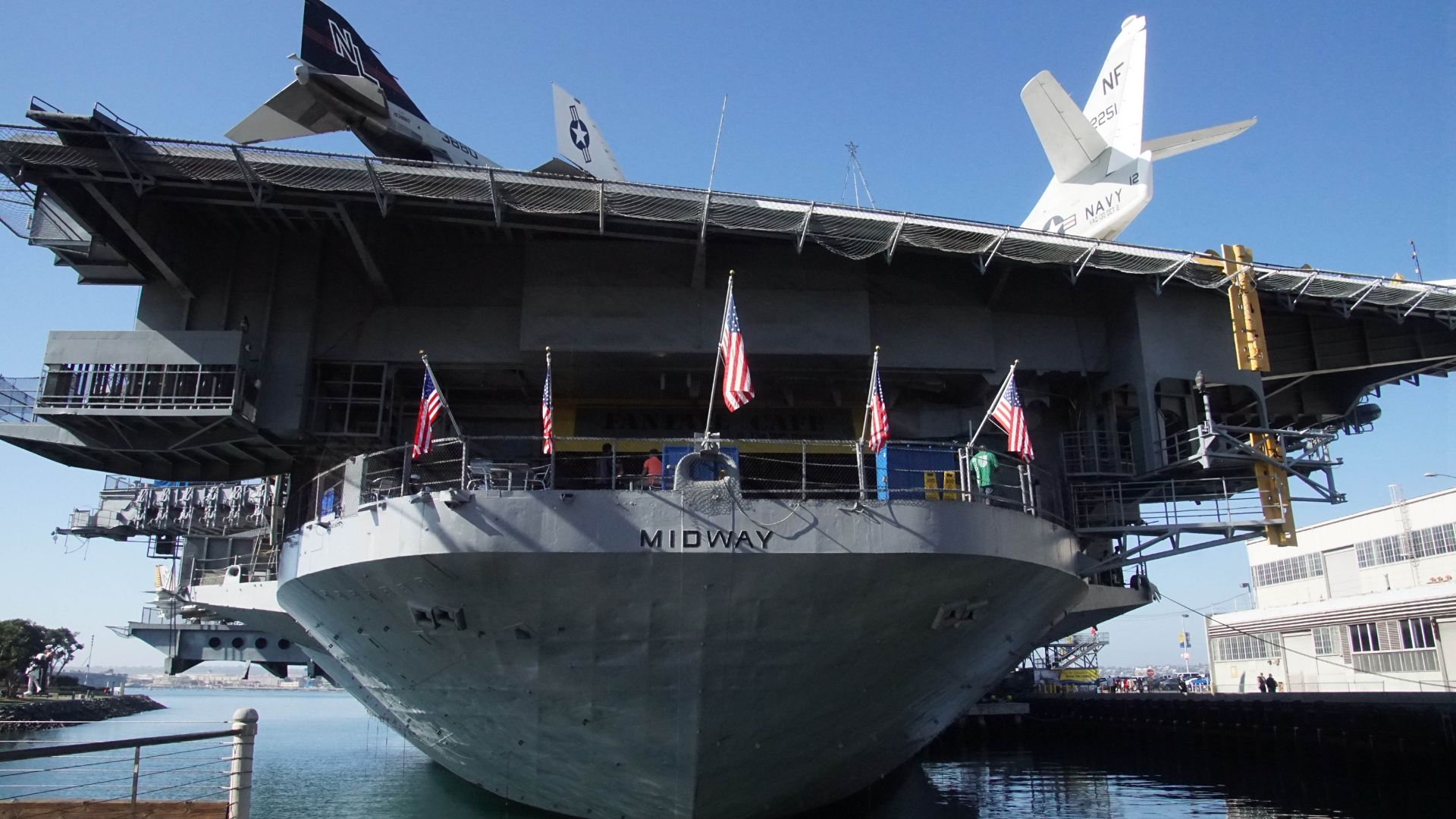 Exterior view of the USS Midway Museum, a historic aircraft carrier in San Diego, California, with the name 'MIDWAY' visible on its bow, and several American flags flying, under a clear blue sky. Two aircraft are visible on the flight deck.