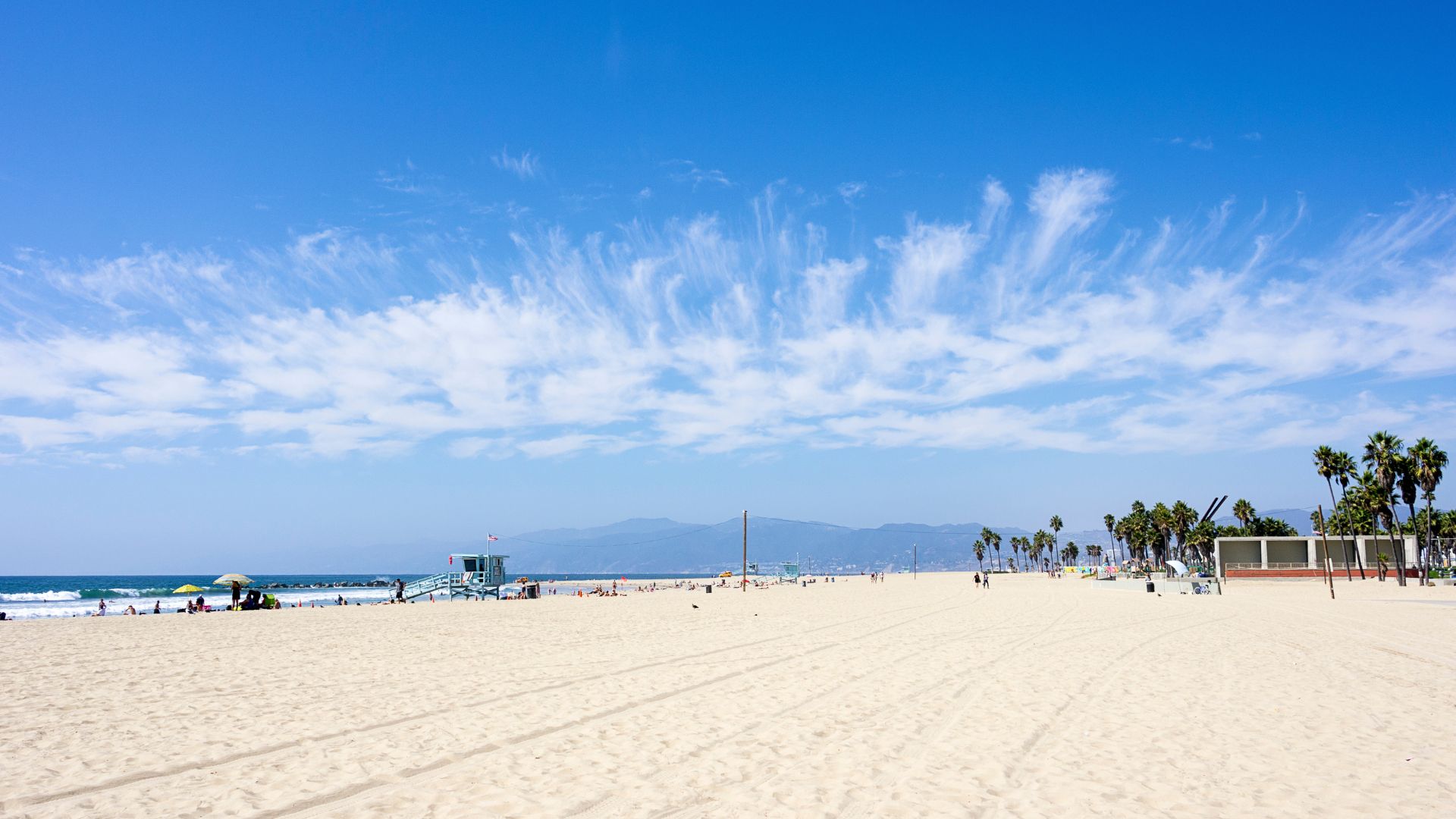 Venice Beach Los Angeles California, wide shot of sandy beach, lifeguard tower, volleyball courts, palm trees, and mountains in the distance under a blue sky with wispy clouds, ideal for travel and tourism content.