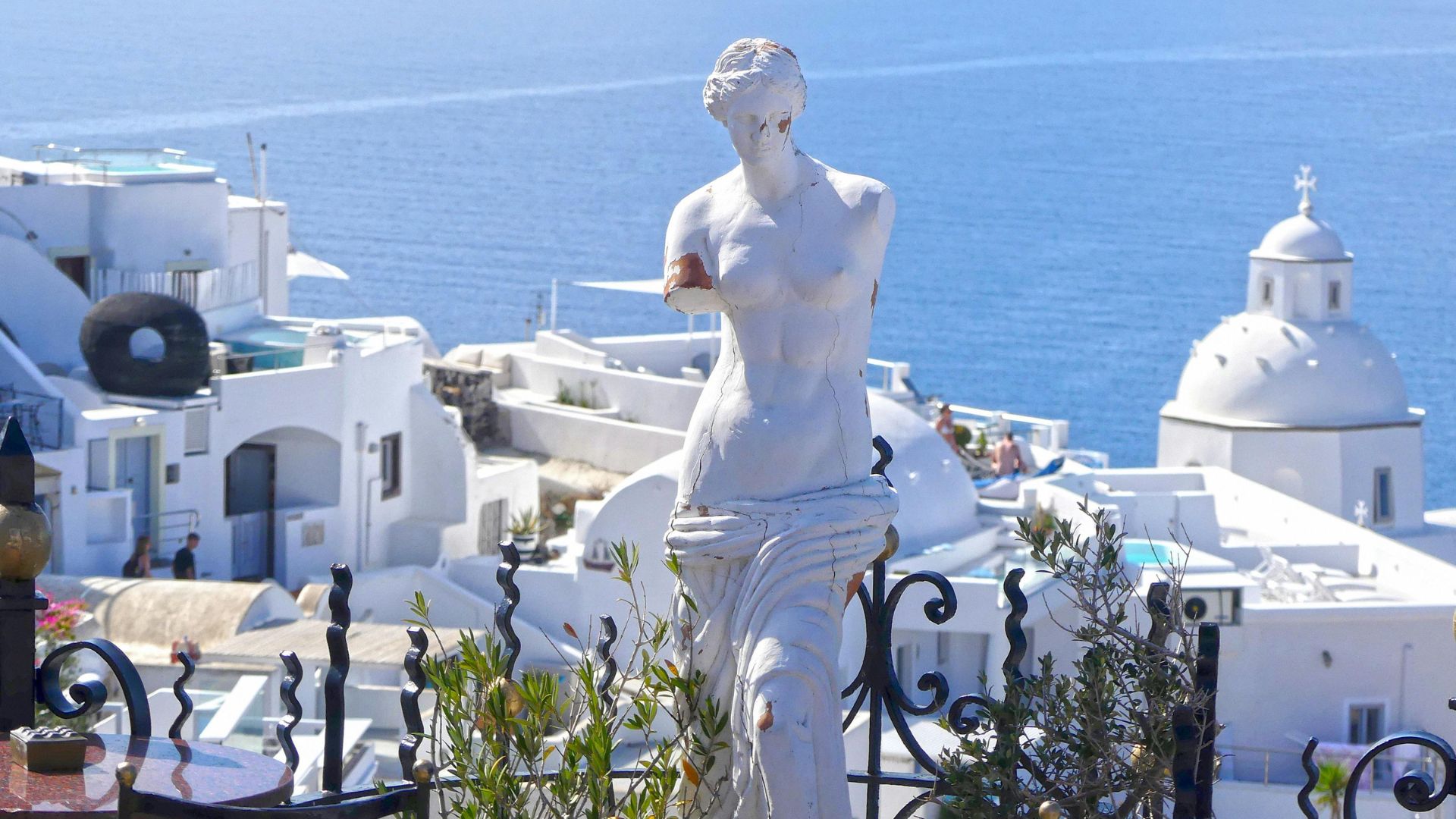 A white replica of the Venus de Milo statue stands overlooking a scenic Greek landscape with whitewashed buildings and blue sea.