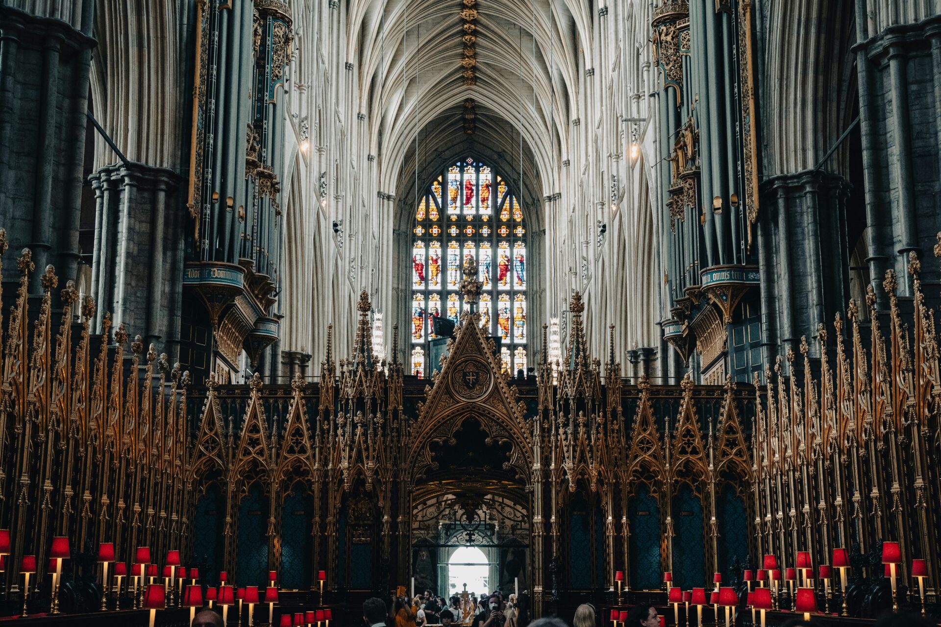 View down the central aisle of Westminster Abbey, showing rows of wooden choir stalls, ornate gothic arches, and a distant glimpse of the high altar under vaulted ceilings.