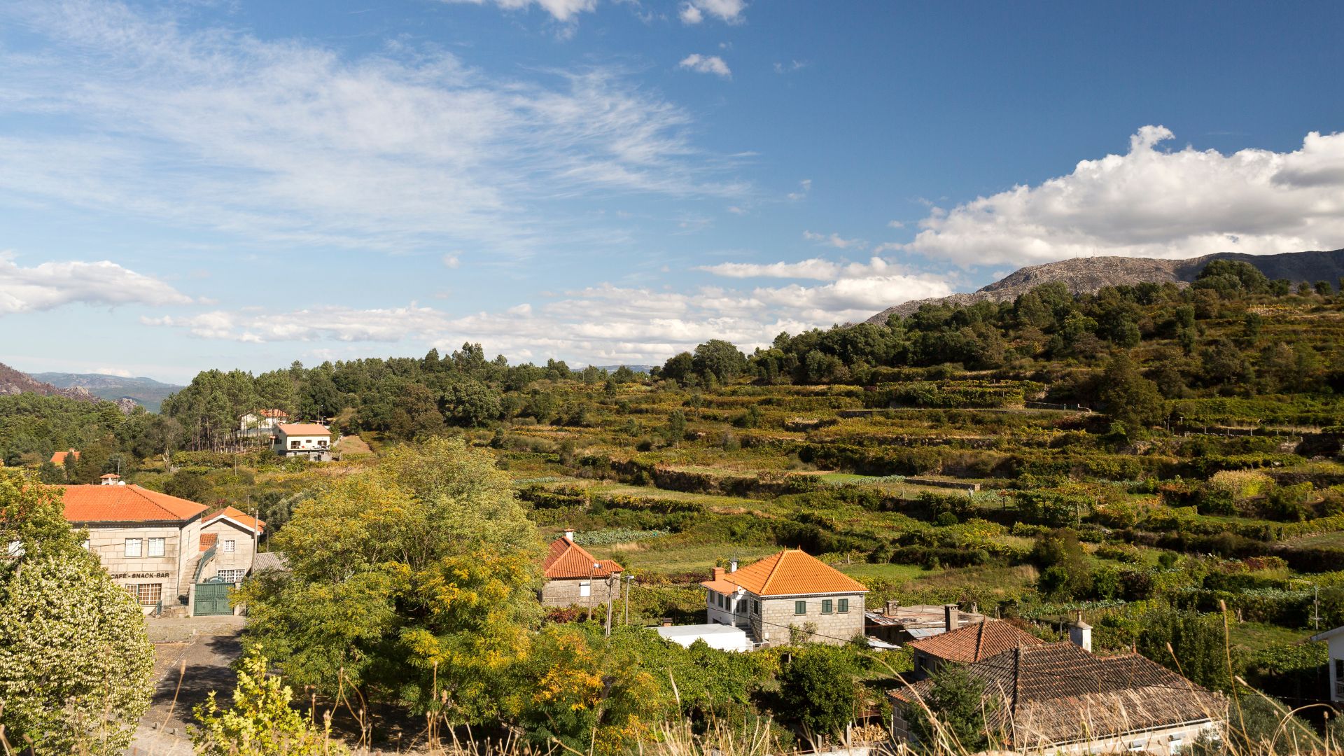 A panoramic view of the traditional Portuguese village of Lindoso, featuring houses with orange-tiled roofs nestled amidst terraced agricultural fields and lush green hills under a partly cloudy sky. 
