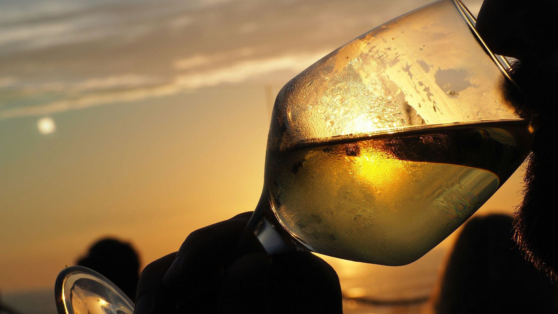 A person holds a glass of white wine against the backdrop of a sunset over the ocean. The sun's reflection shimmers on the water and through the glass, which appears to have condensation on its exterior.