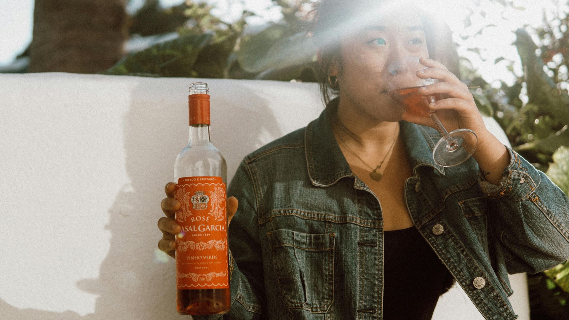 A person in a denim jacket sips rosé wine from a glass while holding a bottle of Casal Garcia Vinho Verde outdoors in sunlight.