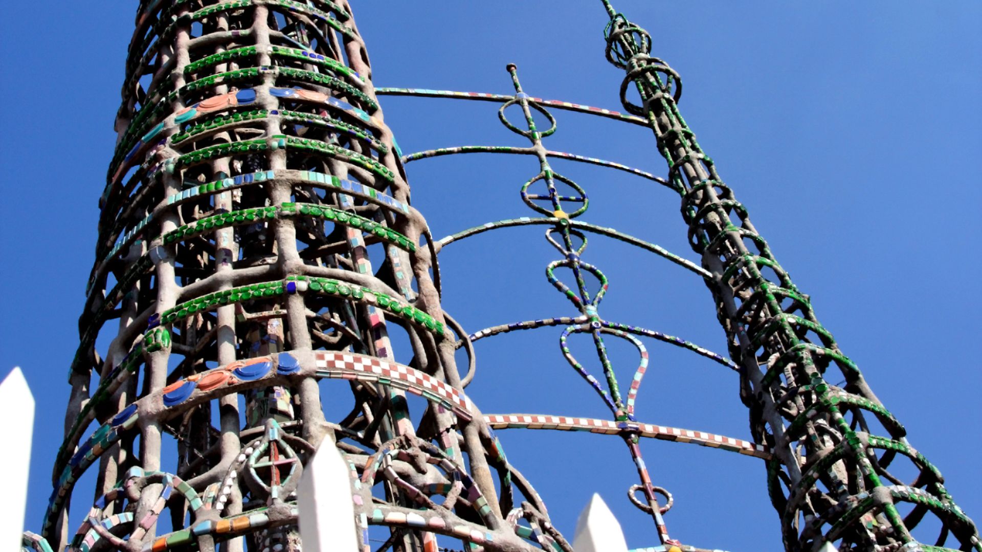A low-angle shot of the Watts Towers, showcasing their intricate, open-work construction with colorful accents against a clear blue sky. The towers are made of steel and concrete, adorned with mosaics of glass, tile, and shells, and are partially visible behind a white picket fence.