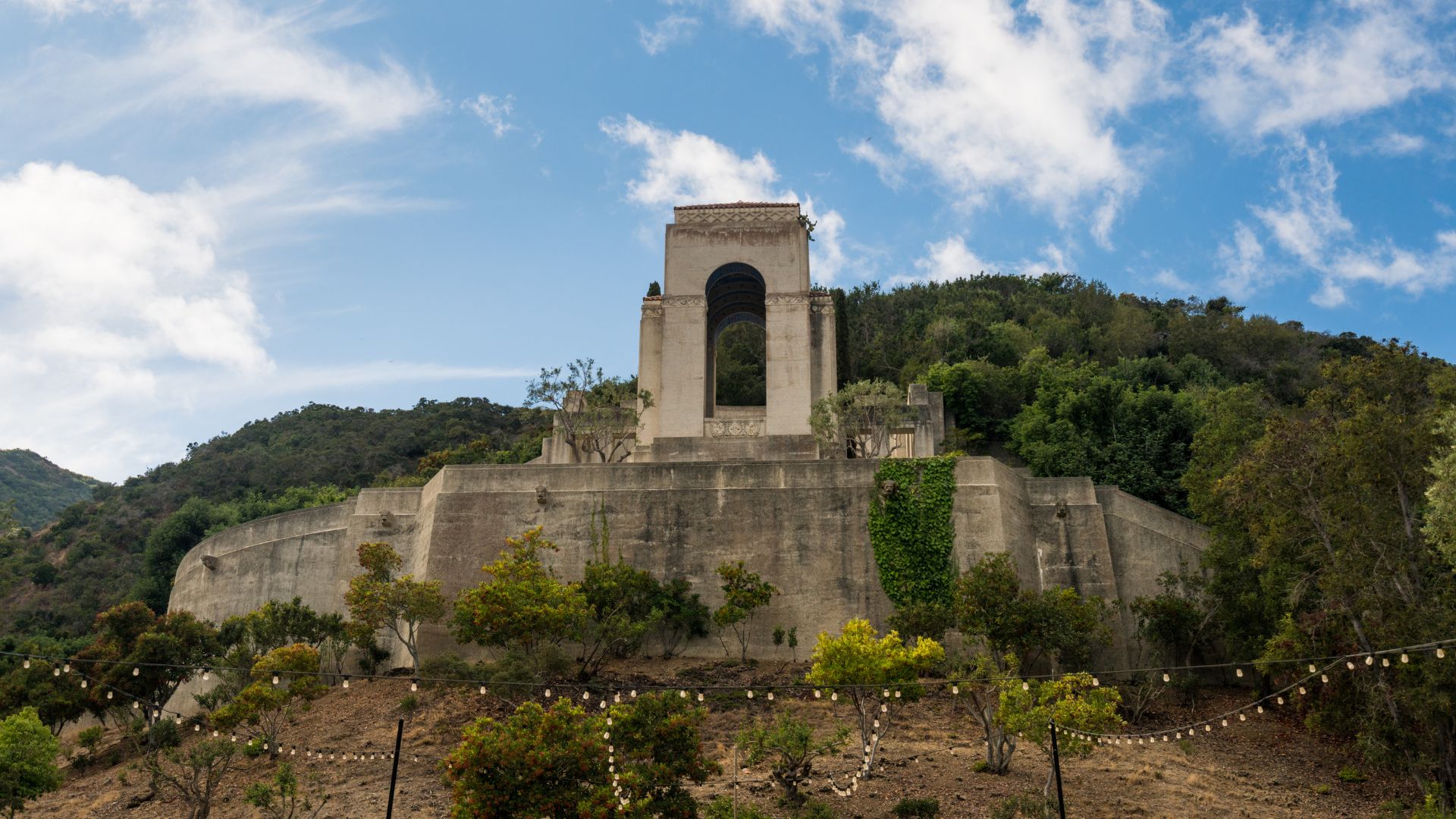 A low-angle shot of the Wrigley Memorial, a large, light-colored stone structure with a prominent arched opening, situated atop a terraced hillside covered in lush green trees and plants. A string of lights is visible in the foreground, draped across the lower part of the hillside. The sky above is partly cloudy with patches of blue.