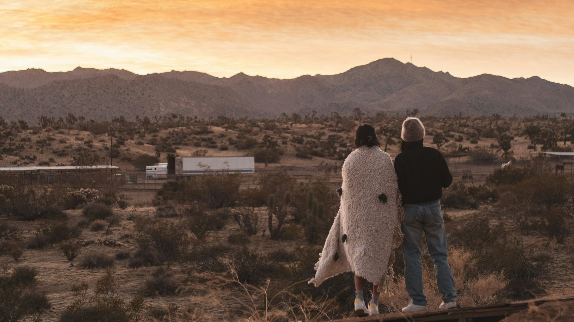 Two people, one wrapped in a blanket, stand on a small wooden platform overlooking a vast desert landscape at sunset. In the distance, mountains rise under an orange and yellow sky, with scattered desert vegetation and a few structures visible in the midground.
