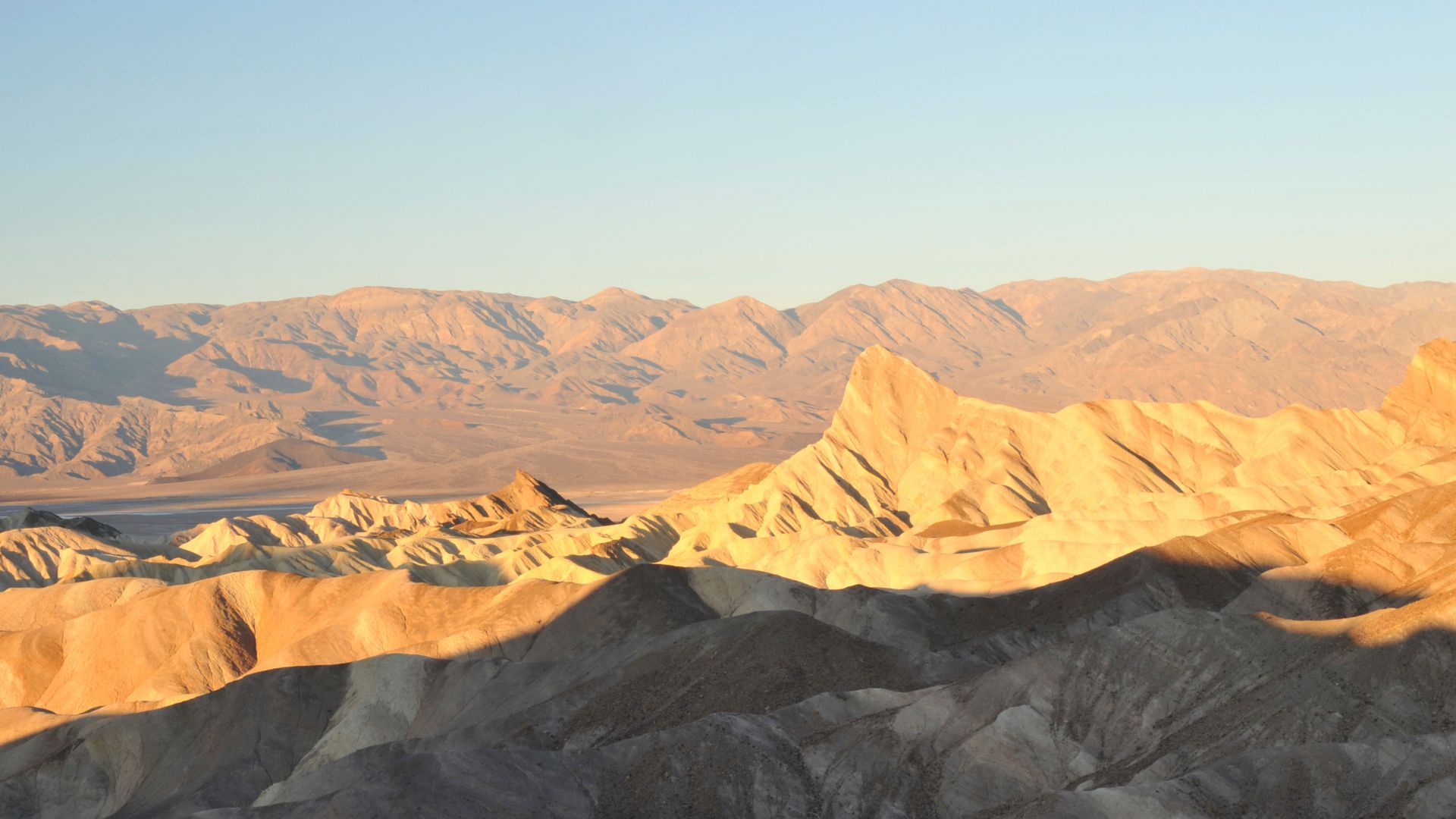 A wide-angle landscape view of Zabriskie Point in Death Valley National Park, showing undulating golden-colored badlands in the foreground and midground under a clear sky.