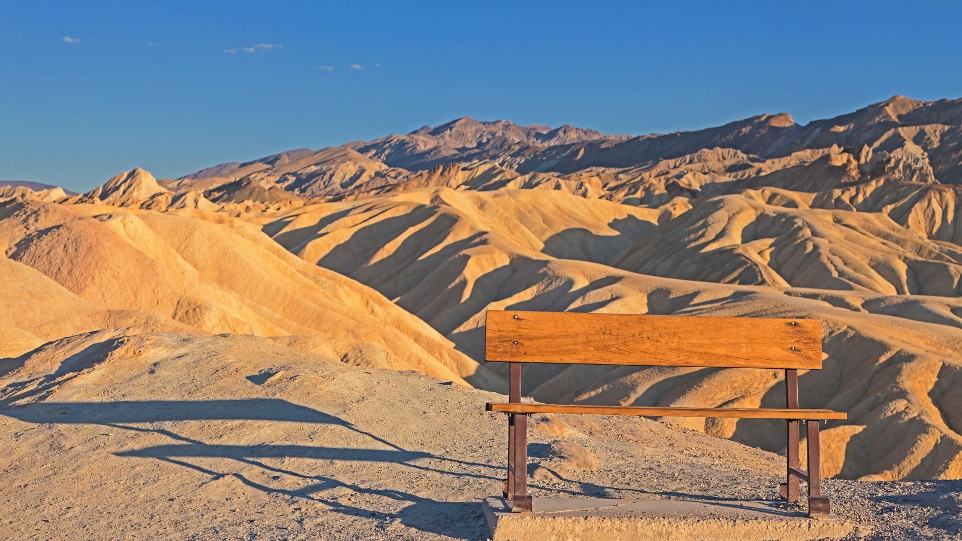 A wide-angle landscape view of Zabriskie Point in Death Valley National Park, showing undulating golden-colored badlands in the foreground and midground under a clear sky and a bench.