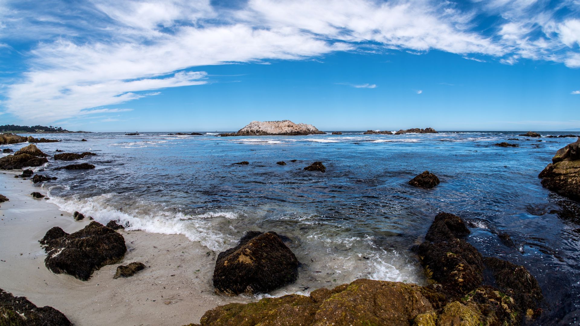 A wide-angle shot of a rocky coastline with waves crashing on a sandy beach, featuring several large rocks in the foreground and a prominent rocky island in the distance under a blue sky with white clouds.