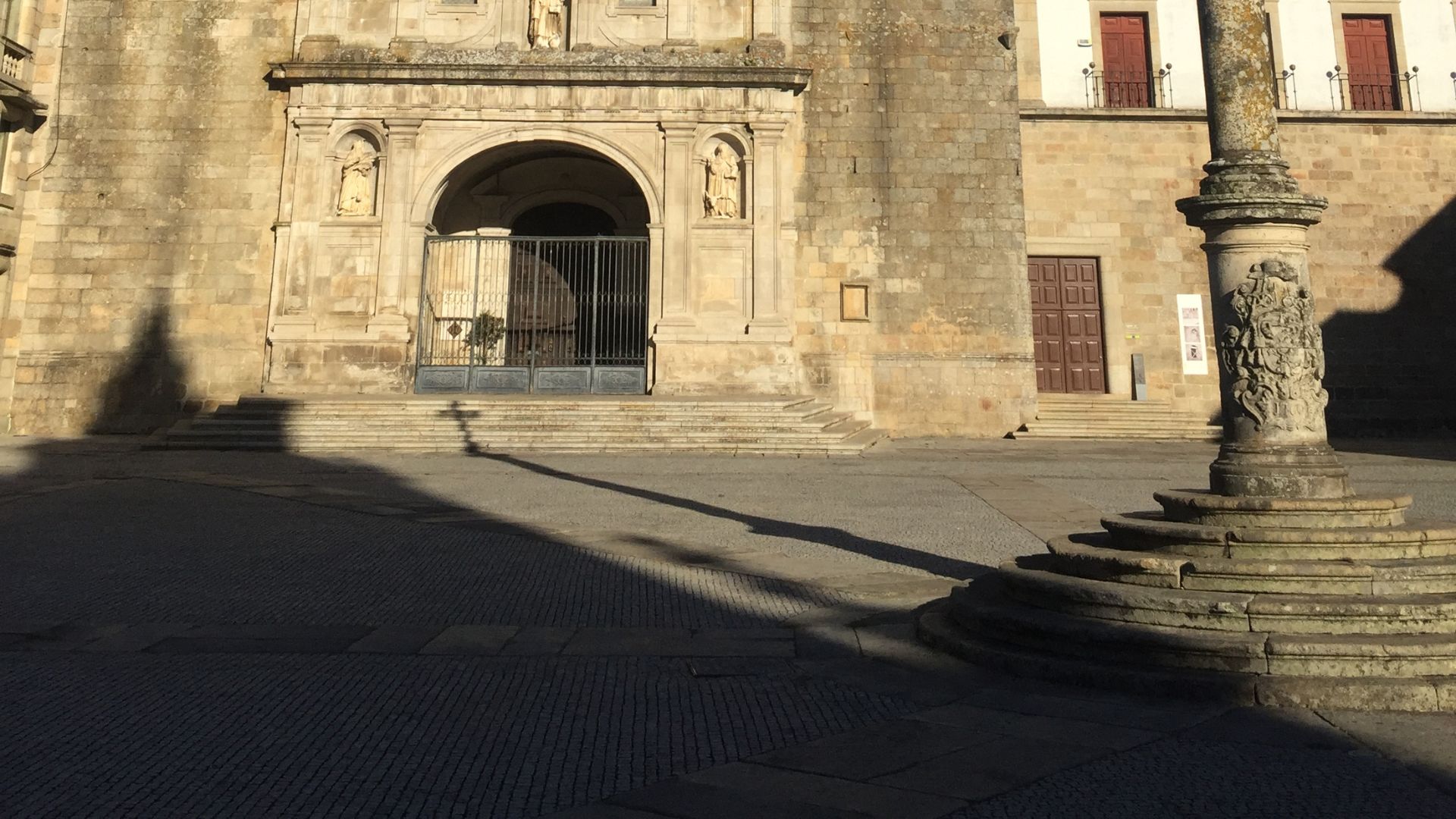 A low-angle shot of the Viseu Cathedral's facade with a stone pillar and its base in the foreground, casting long shadows across the cobblestone square in the late afternoon sun.