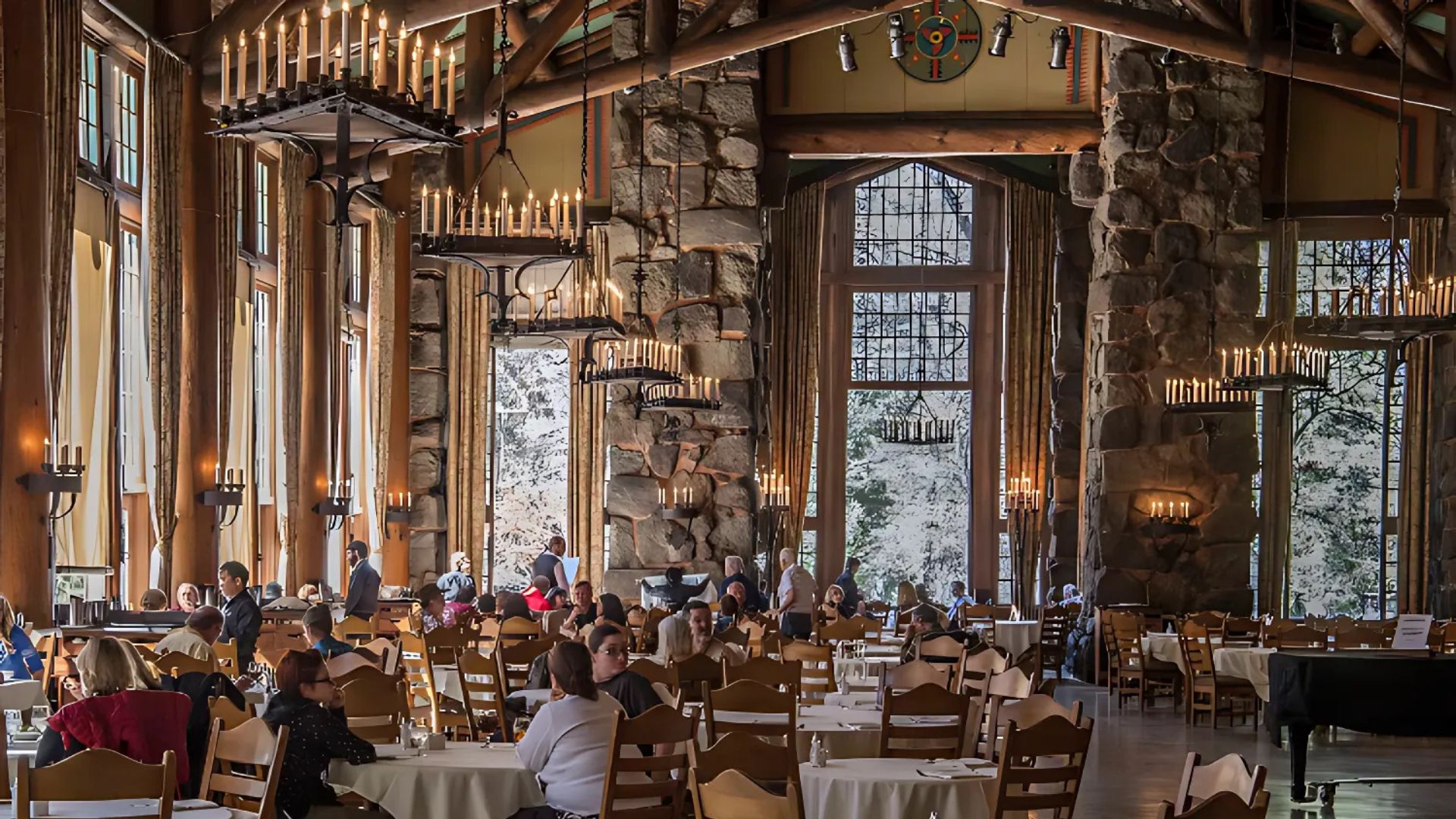 A grand dining hall with high ceilings, large windows, and numerous chandeliers, filled with people seated at tables, suggesting a busy restaurant or hotel dining room. The room features rustic architecture with prominent stone pillars and wooden beams, and large windows offer views of a natural outdoor setting.