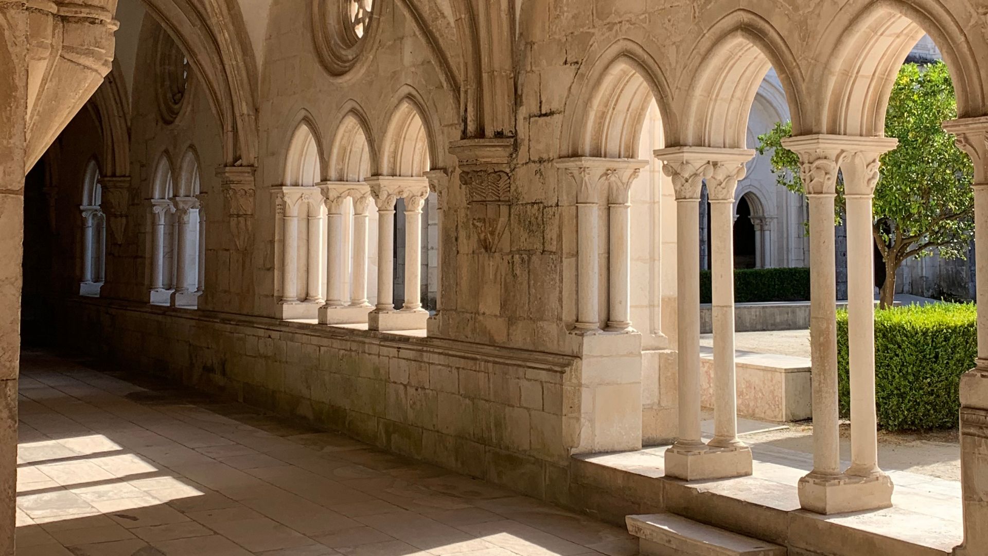A long, arched corridor with stone walls and columns, leading through a historic monastery with sunlight casting shadows on the floor and a courtyard with greenery visible through the arches on the right.