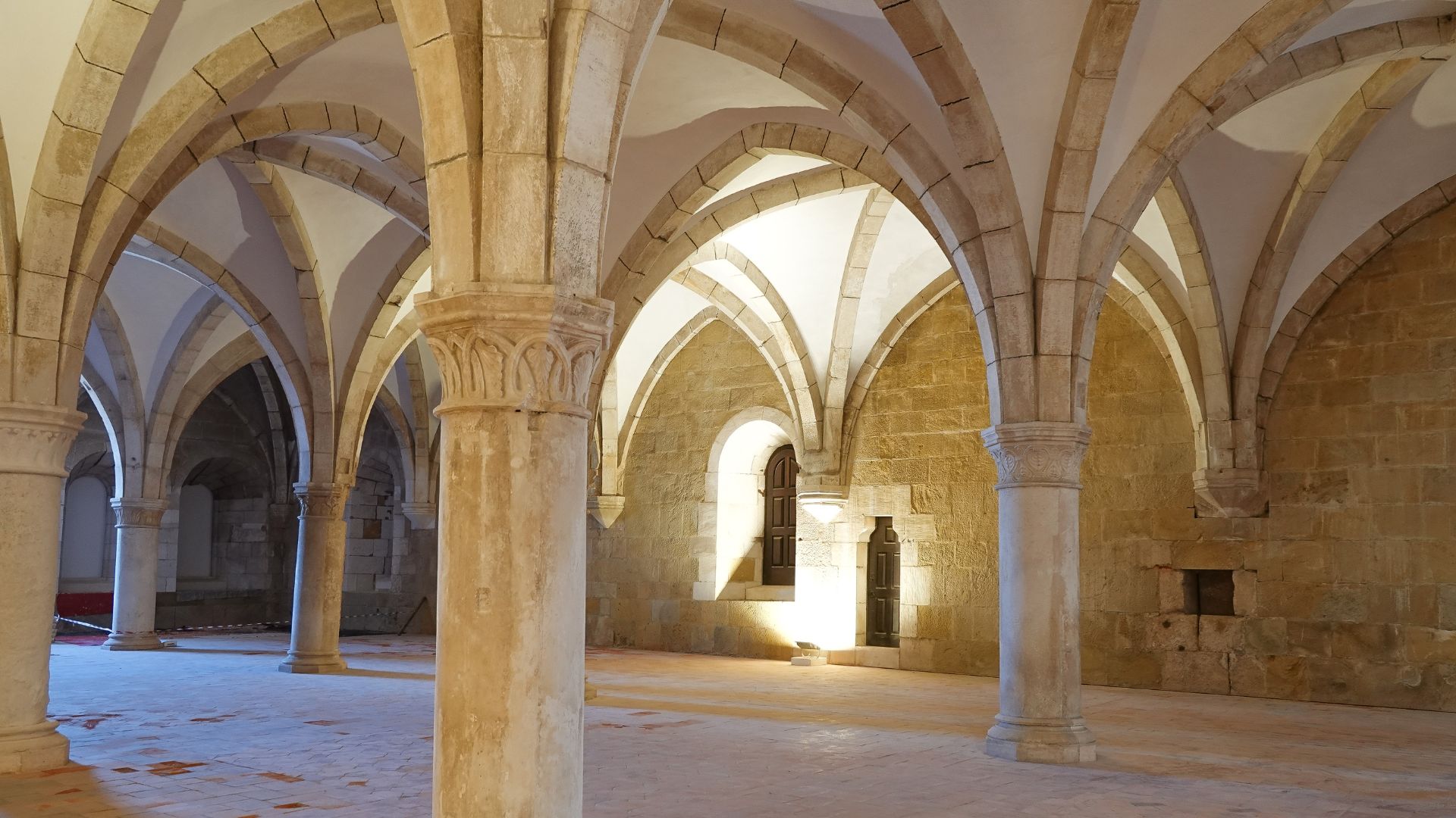 A large, stone hall with numerous thick columns supporting a series of vaulted ceilings, illuminated by light entering through a distant window.