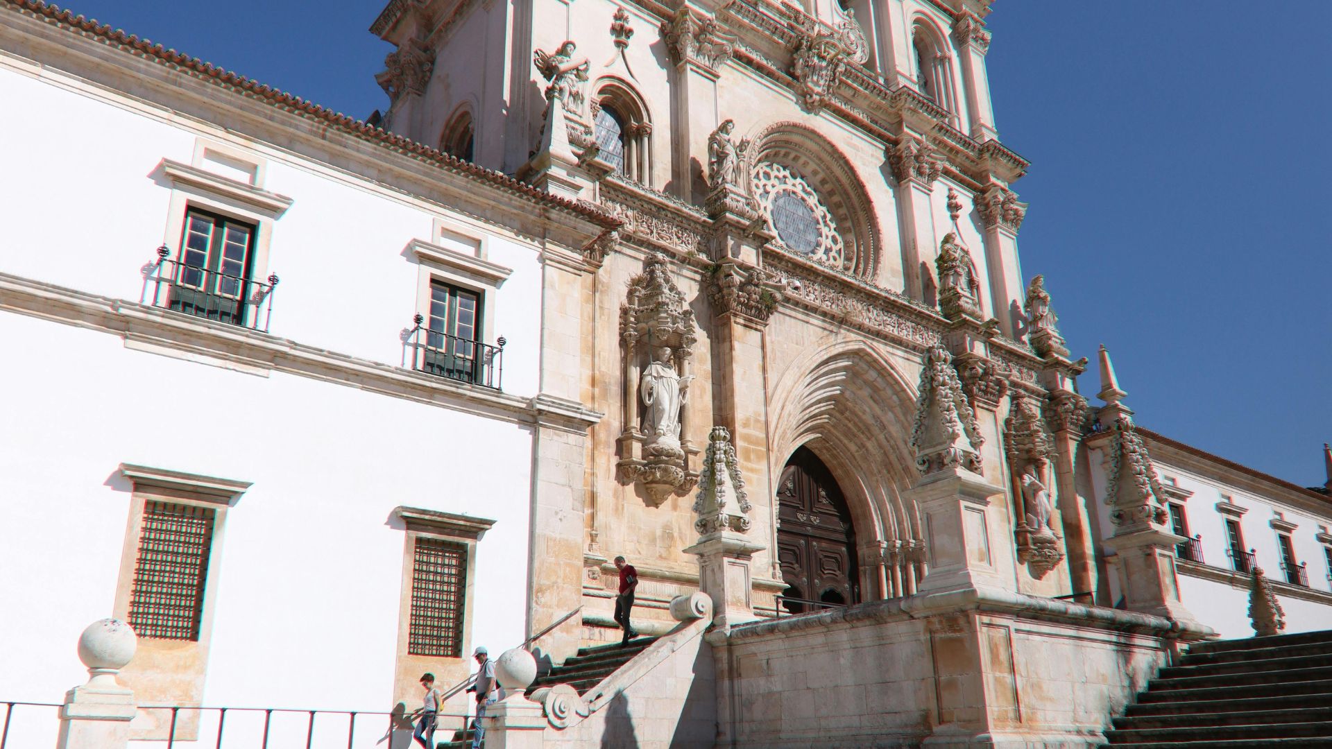 A wide shot of the grand, white facade of the Alcobaça Monastery in Portugal.  The image captures the ornate entrance and part of the adjacent monastic buildings under a clear blue sky, with a few people visible on the steps leading to the entrance. 