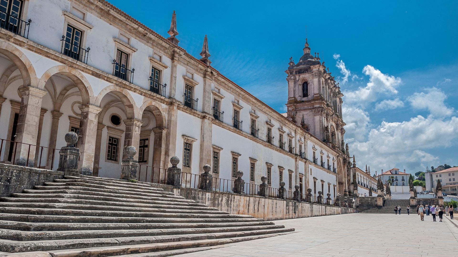 A low-angle view of the grand, white stone Alcobaça Monastery in Portugal, featuring a wide staircase leading up to its imposing facade with arched windows and a prominent tower under a clear blue sky with scattered clouds.