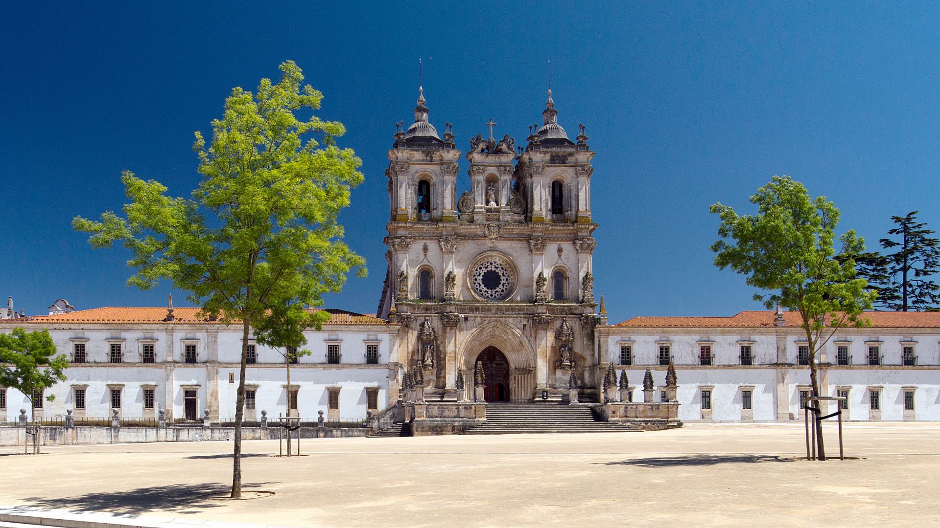 A wide shot of the Alcobaça Monastery in central Portugal, featuring its grand, ornate Gothic facade with two prominent bell towers under a clear blue sky, flanked by white monastic buildings and a sparsely treed, open sandy plaza in the foreground.