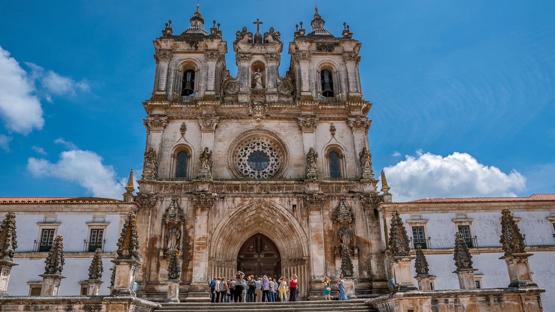 A grand, ornate stone monastery with a large central facade featuring a prominent rose window, two bell towers, and a large arched entrance, with a group of people gathered at the base of the steps on a sunny day under a blue sky with some clouds.