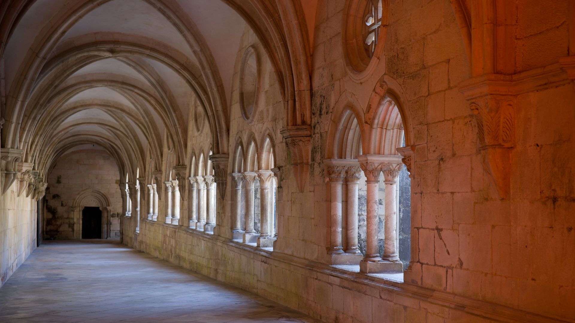 A long, arched stone hallway within a historic monastery, featuring a series of large arched windows on the right side that let in natural light, illuminating the ancient stone walls and vaulted ceiling.