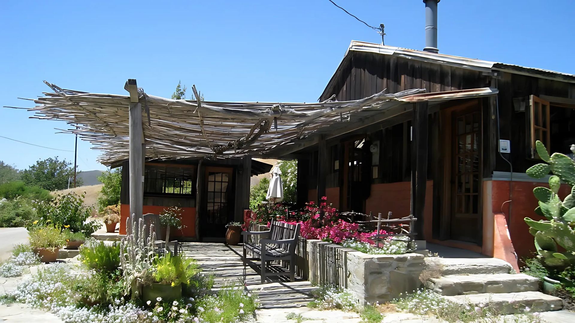 A rustic, wood-sided building with a shaded patio and lush garden area, featuring a stone pathway and steps, under a clear blue sky in a sunny, rural setting.