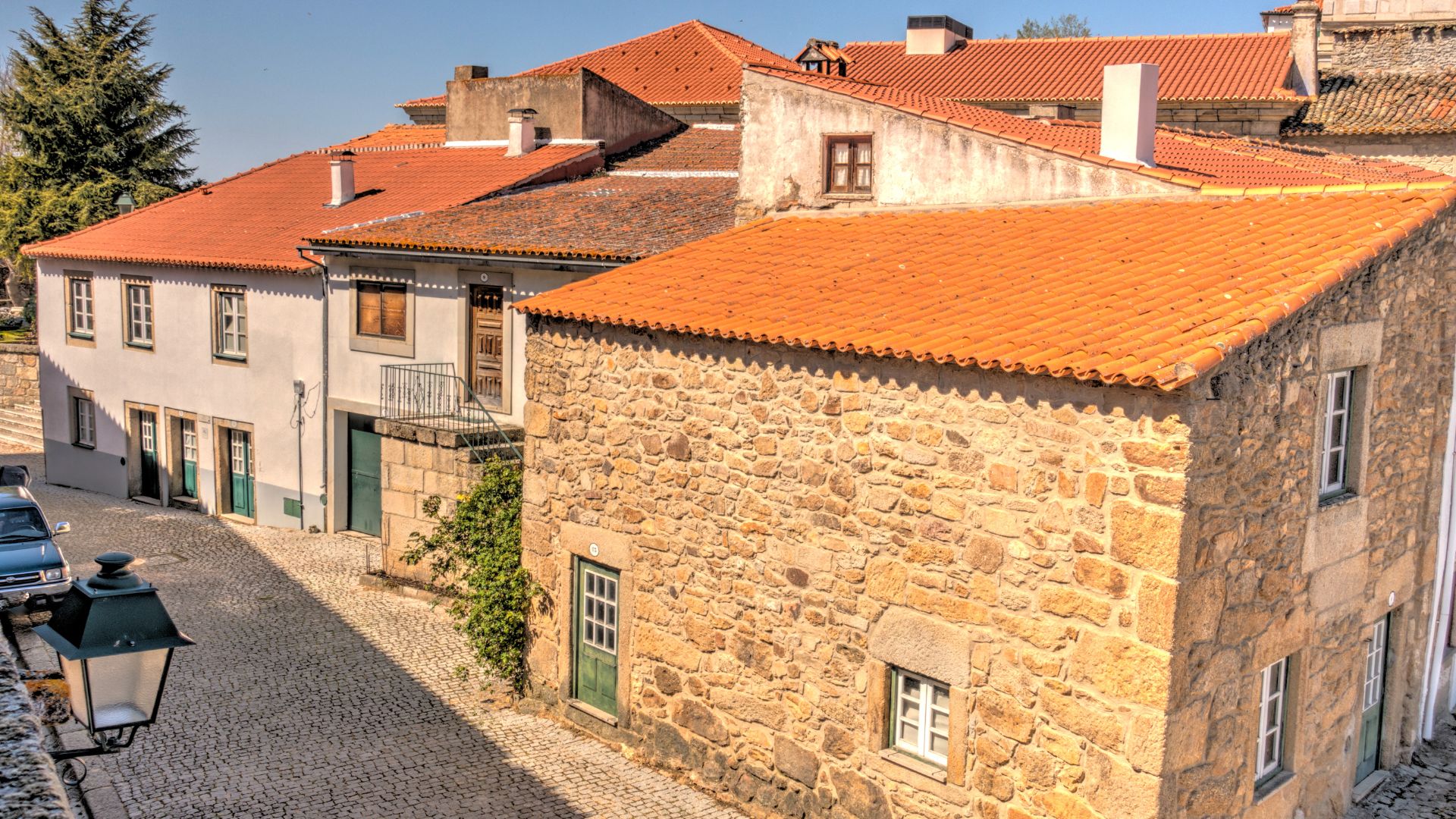 A narrow, sunlit cobblestone street in Almeida, Portugal, featuring historic stone and whitewashed buildings with orange tiled roofs and green doors, leading towards a larger stone structure.